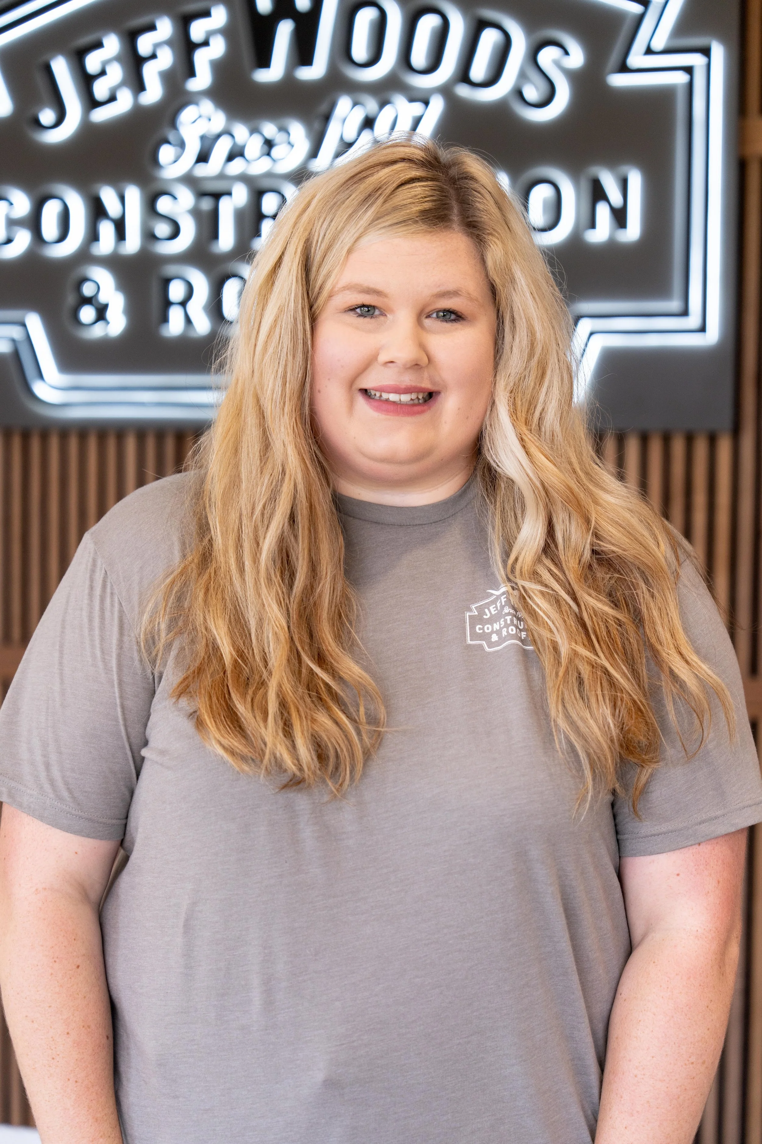 A woman with long, wavy blonde hair wearing a gray t-shirt with a company logo, standing in front of a neon sign for Jeff Woods Construction & Roofing.