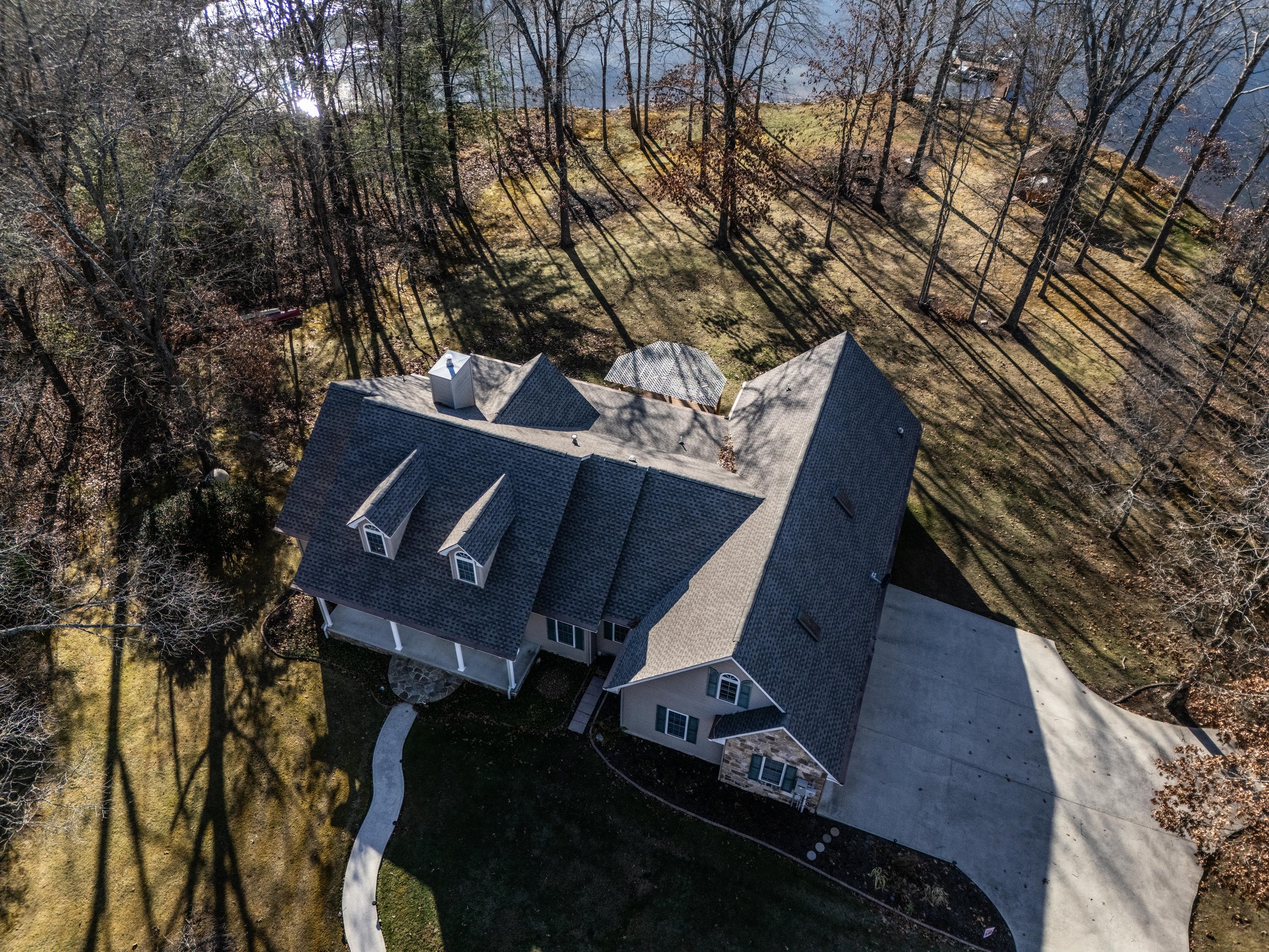 Aerial view of a large house with a gray roof and driveway, surrounded by bare trees and an expansive lawn sloping down to a lake.