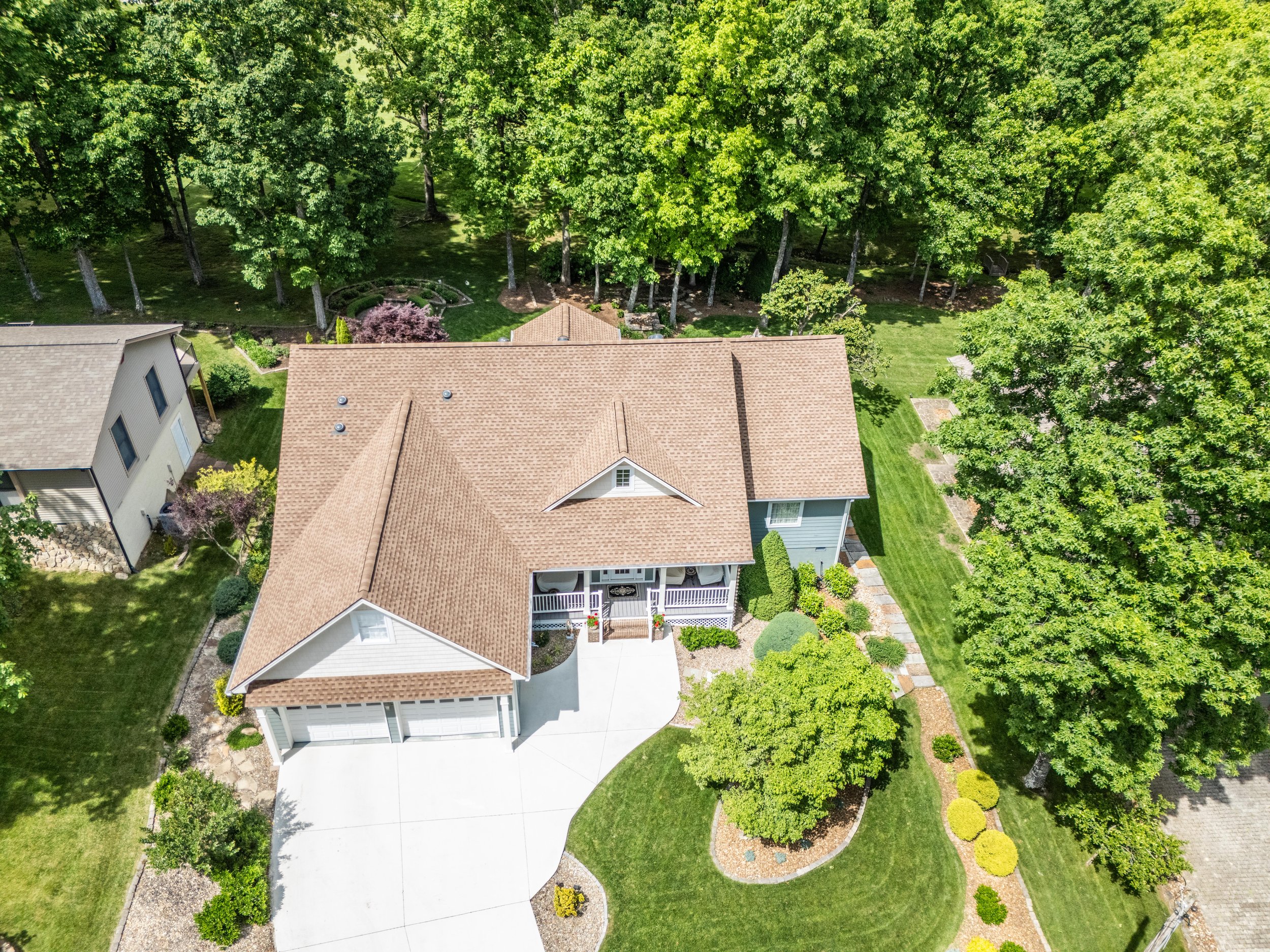 Aerial view of a suburban house with a beige roof, surrounded by a well-maintained lawn and various trees. Includes a driveway leading to a garage and landscaped garden area in the front yard.