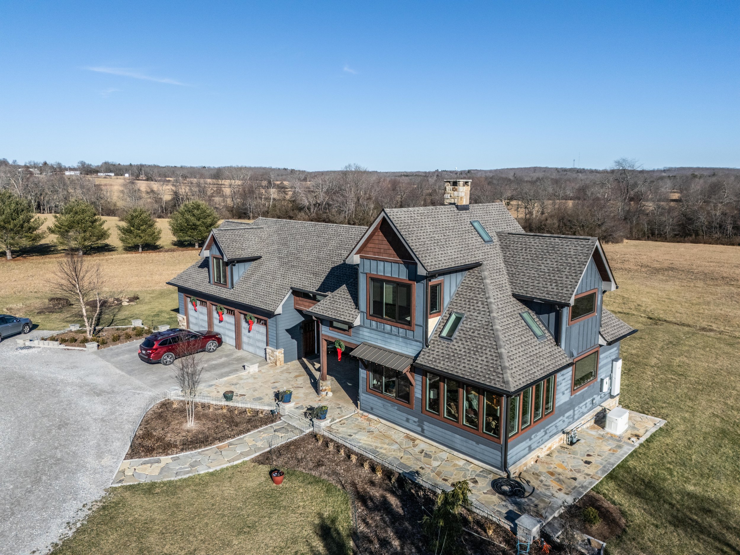 Aerial view of a large modern house in a rural setting with a stone driveway, multiple gables, and a stone chimney. The house is decorated with red ribbons and garlands, indicating a festive holiday season. There are a few cars parked in front and th