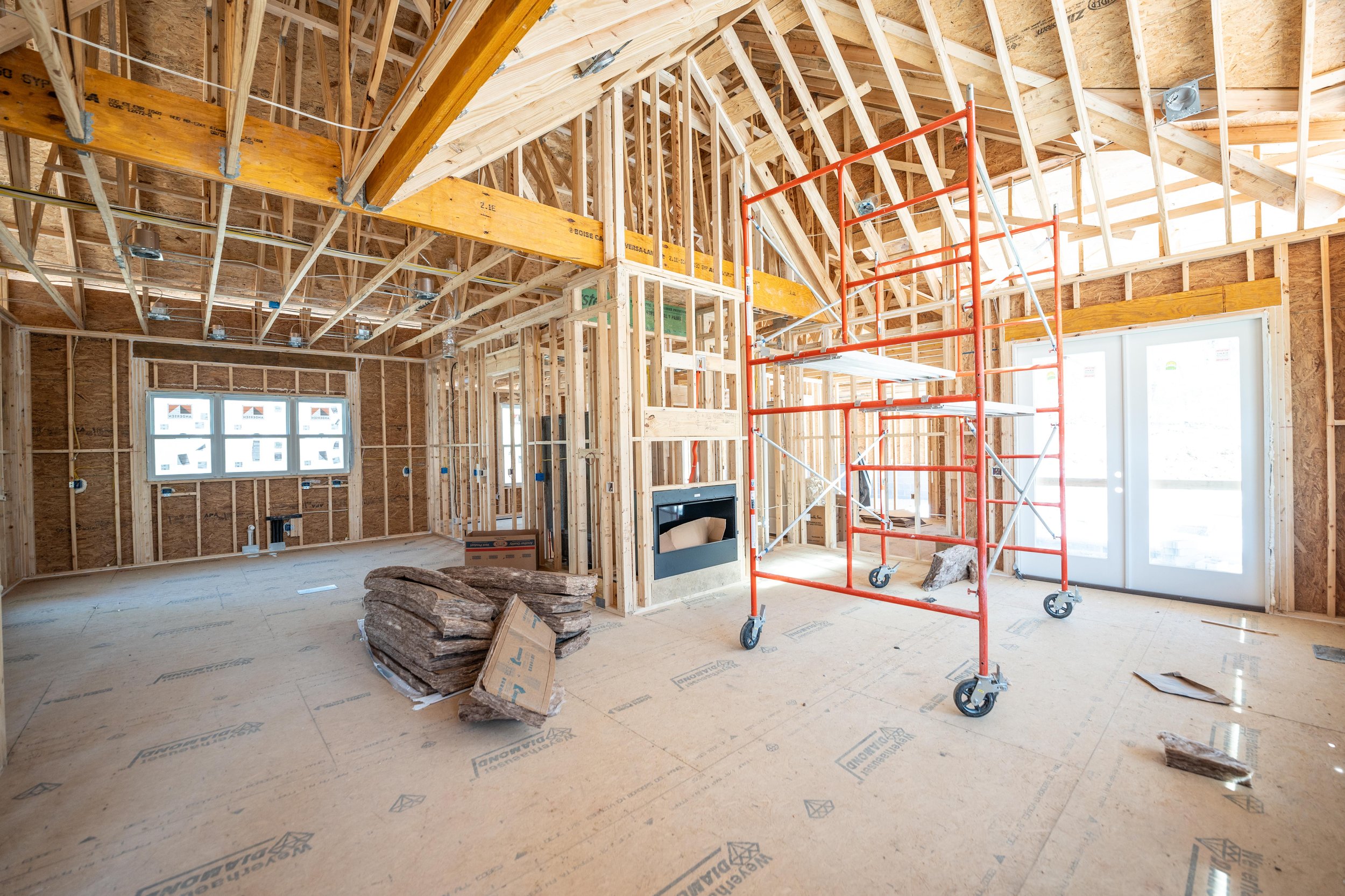 Interior of a house under construction, showing wooden framing, exposed beams, a red metal scaffold, and unfinished walls.