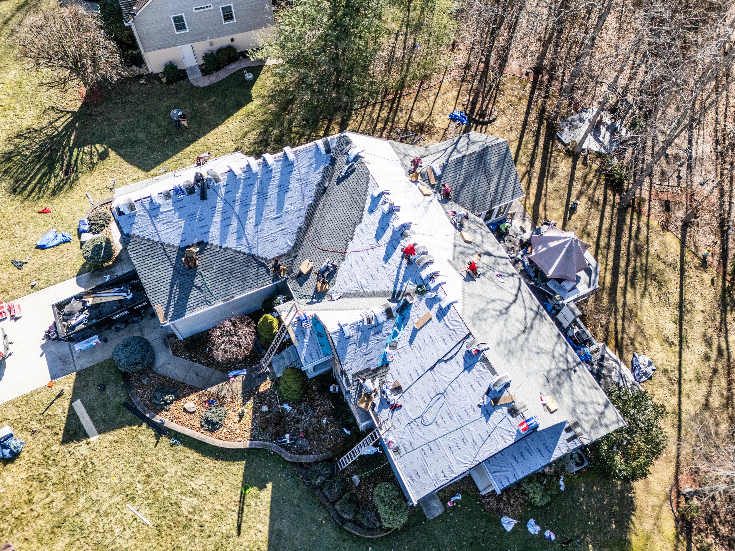 Aerial view of a large house undergoing roof repair, with workers on the rooftop and construction materials scattered around the yard.
