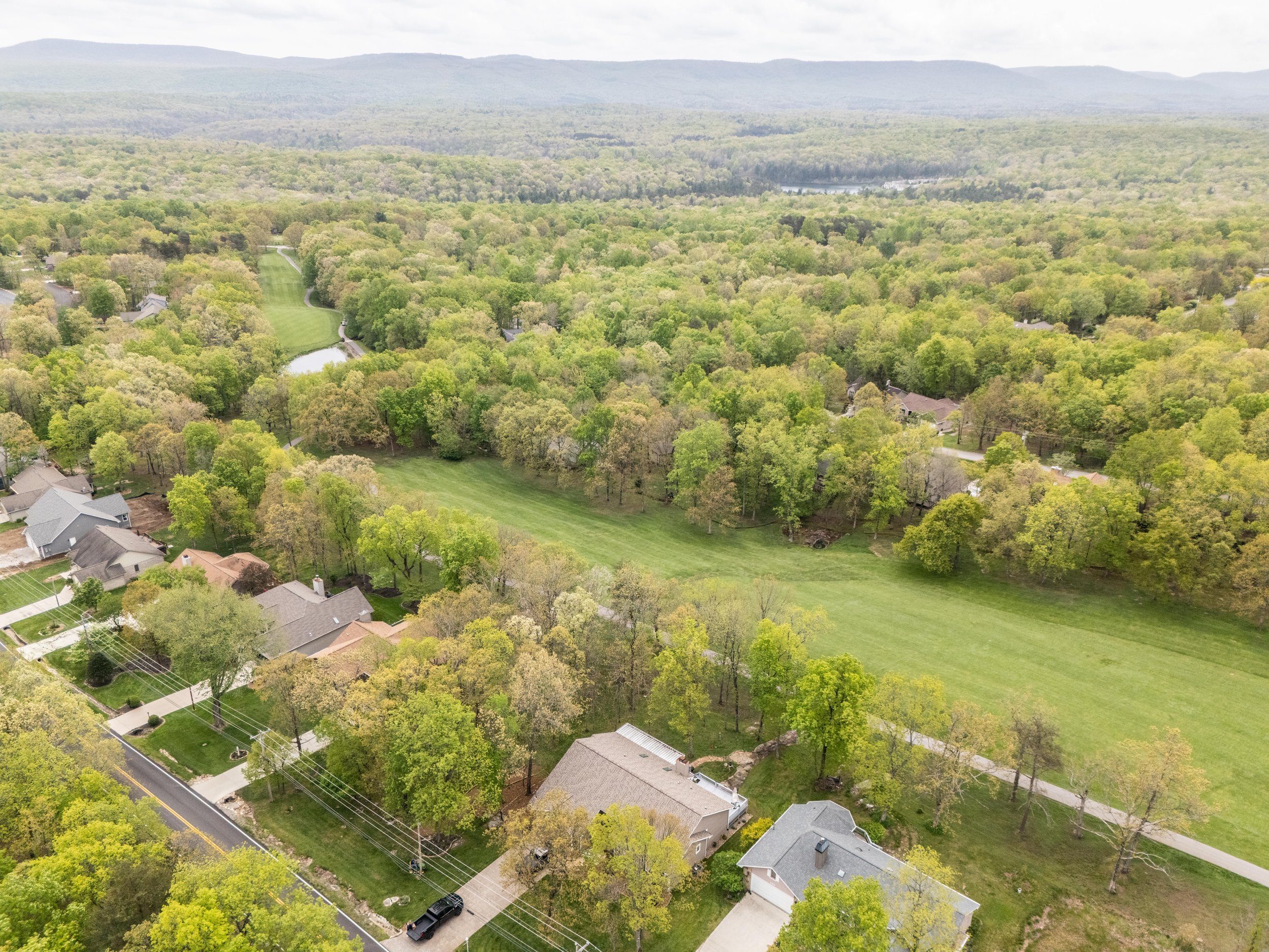 Aerial view of a suburban neighborhood with houses, green lawns, and a road. The area is surrounded by dense forests and rolling hills. A golf course or park area is visible, characterized by open grassy spaces and a winding path. Mountains in the ba