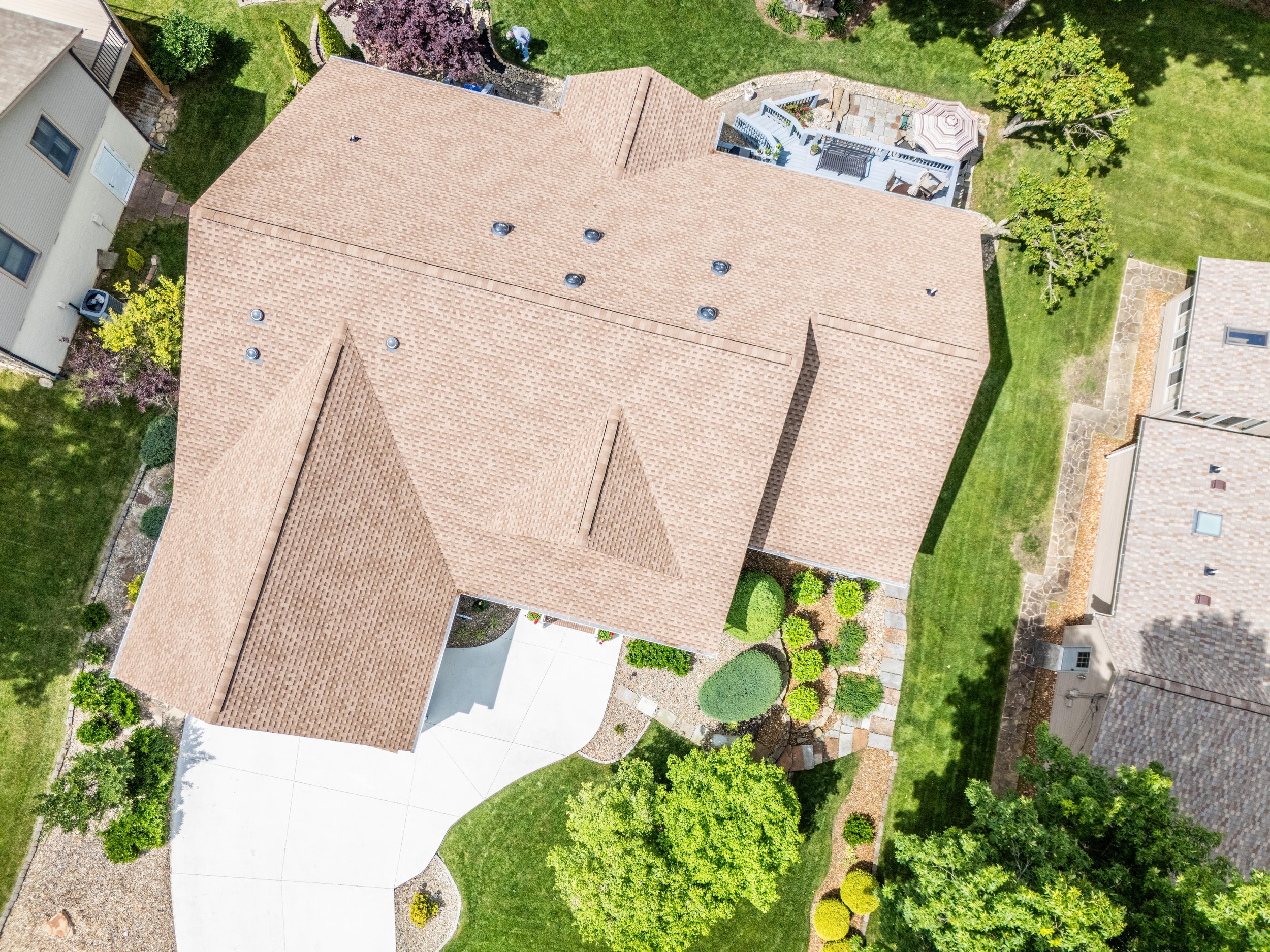 Aerial view of a house with a brown shingled roof, surrounded by a green lawn and garden. The property includes a patio with outdoor furniture and a pathway leading to the driveway. Neighboring houses and landscaped yards are visible on either side.