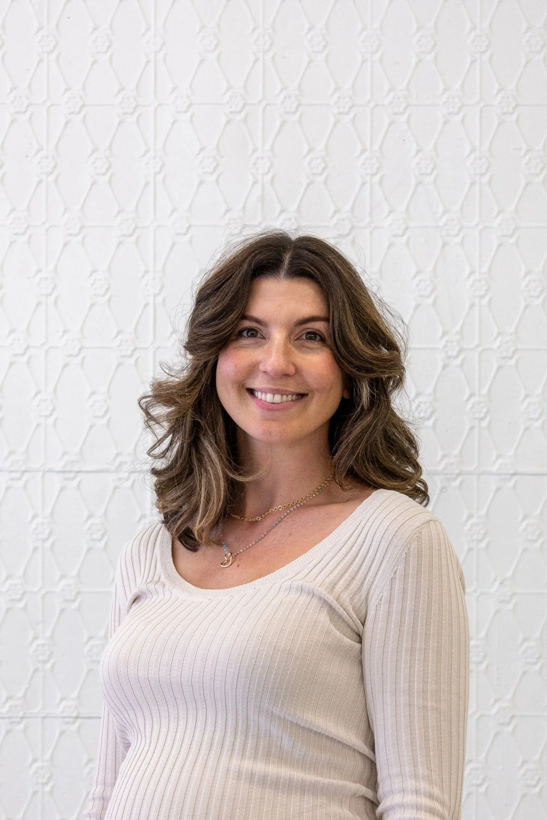 A woman with long brown hair tied to one side, smiling, standing against a beige wall, wearing a white dress with scalloped edges and button details, and a small gold necklace.