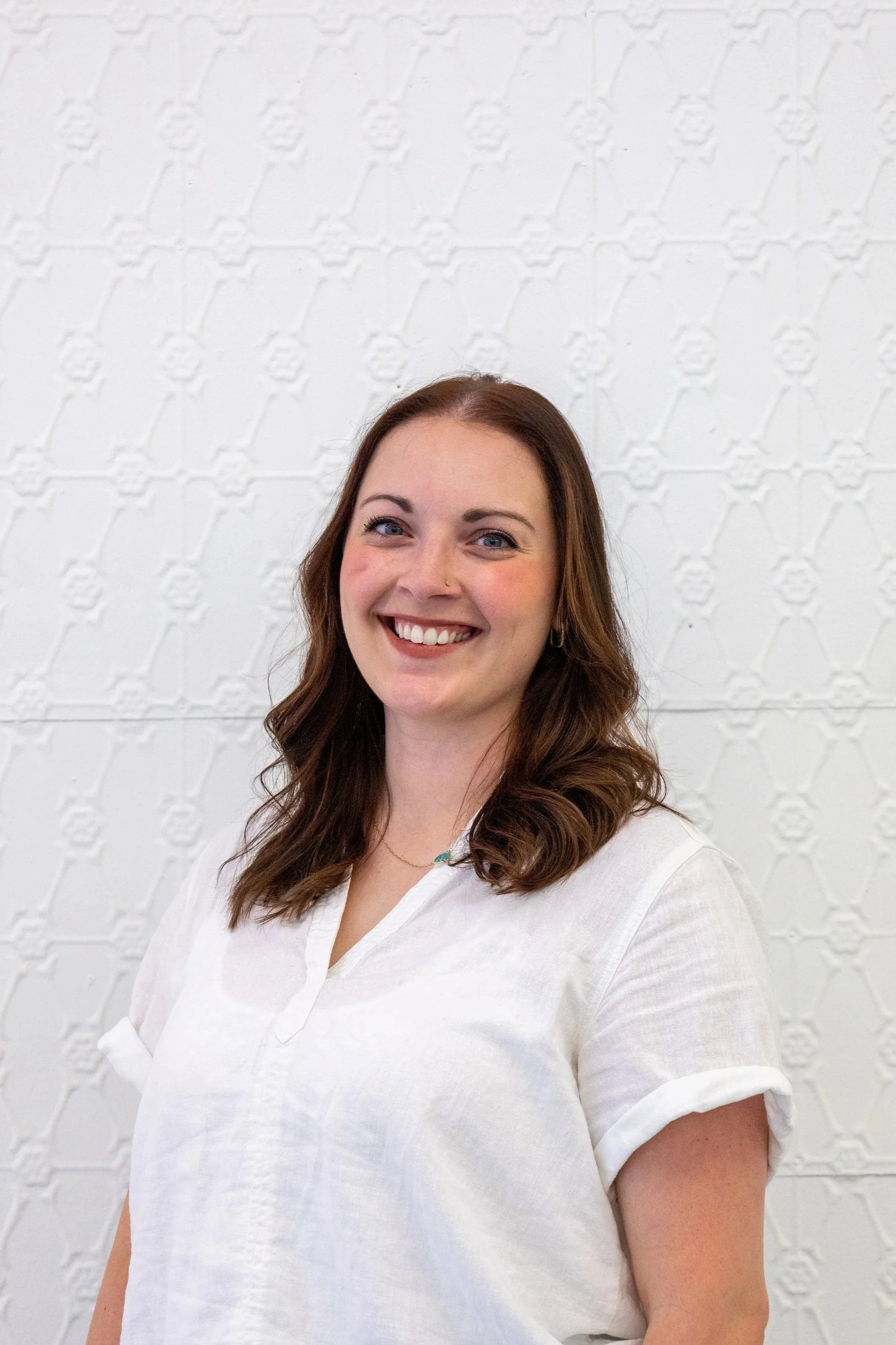 A young woman with shoulder-length dark hair smiling in front of a cream wall with a textured pattern. She is wearing a light-colored, short-sleeved shirt, blue jeans, hoop earrings, a small necklace, and a nose stud.