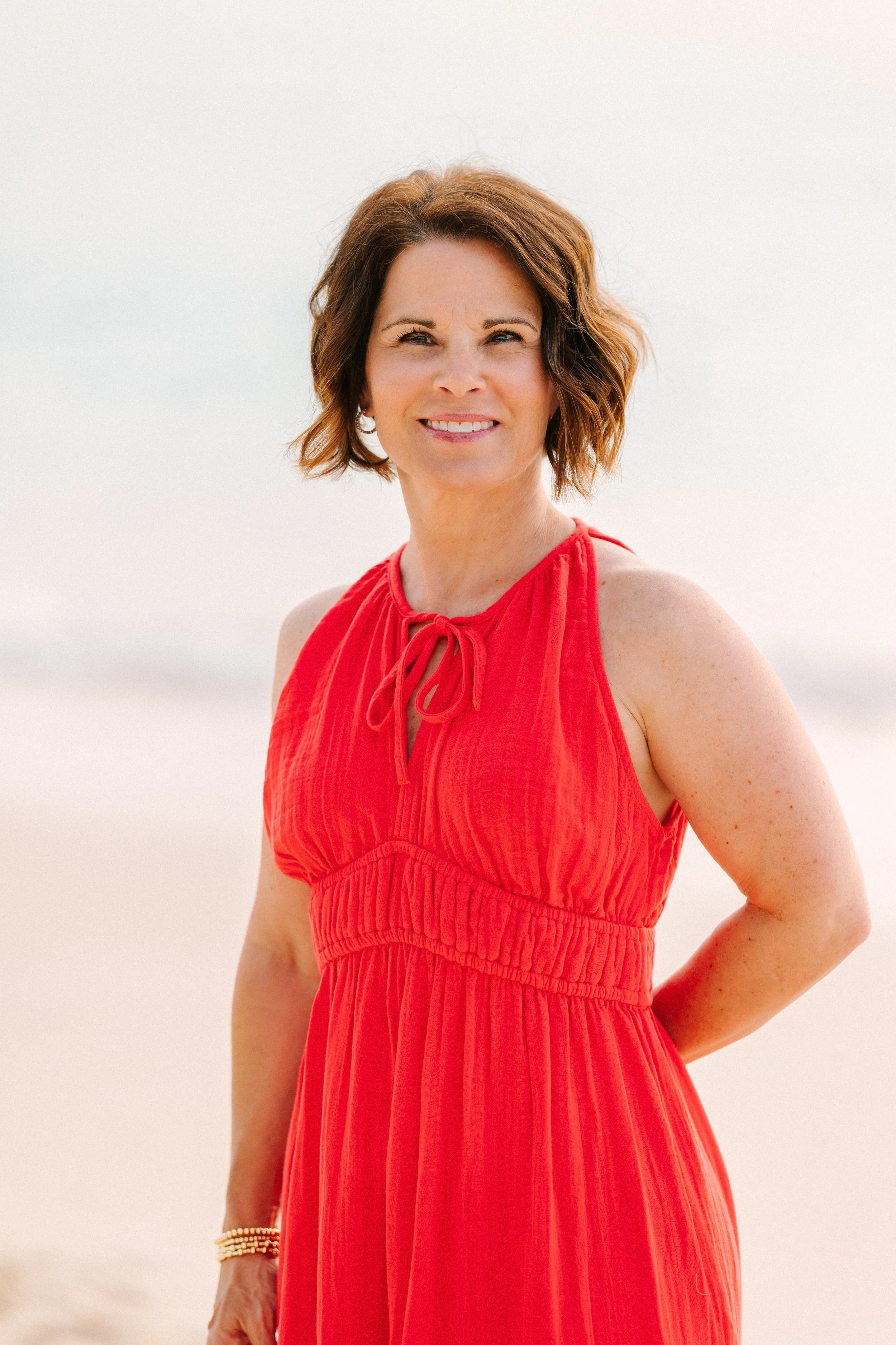 A woman with long, curly brown hair and fair skin, wearing a red sweater, standing against a plain light-colored wall, smiling softly at the camera.