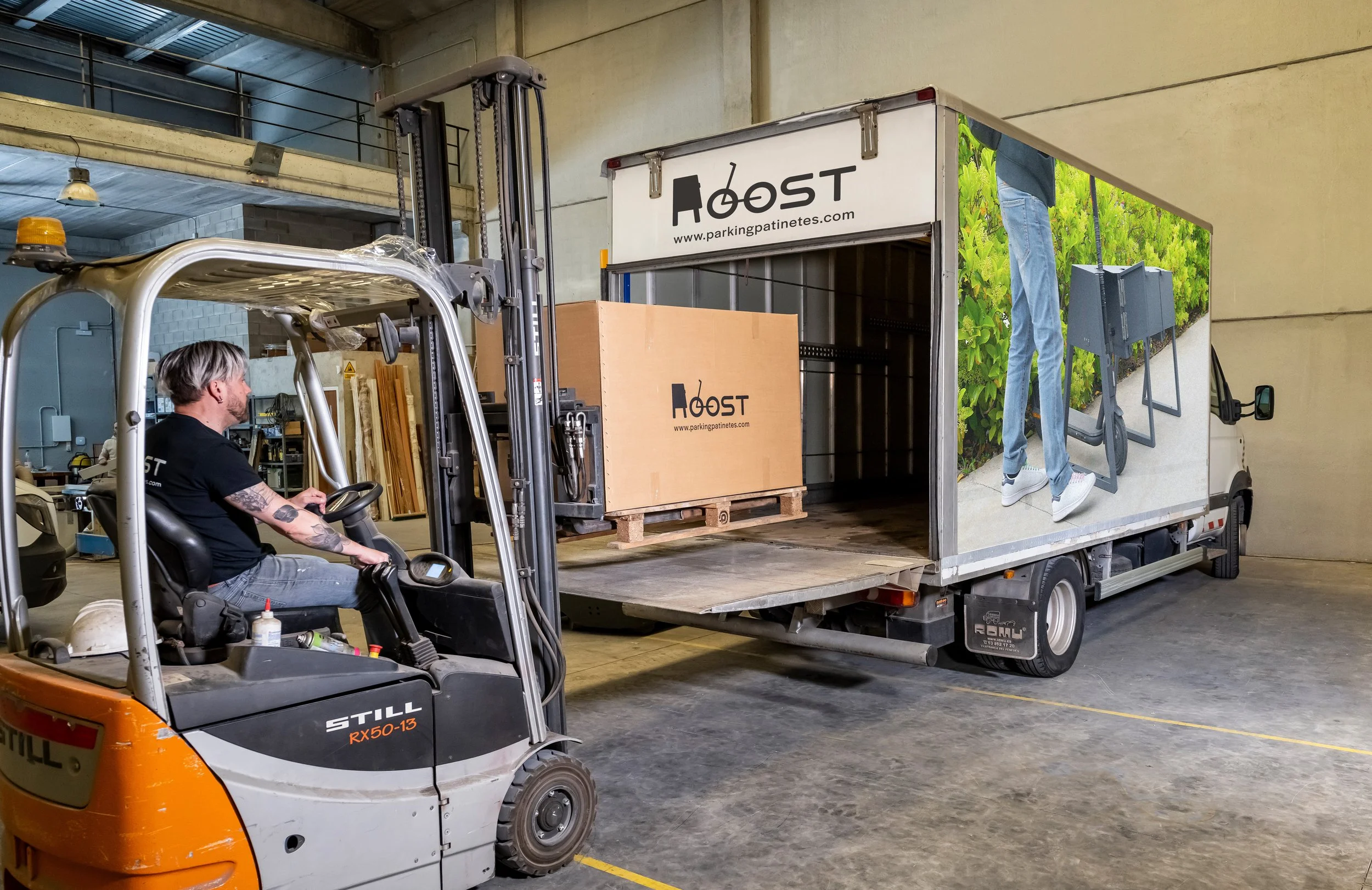Un hombre en un montacargas mueve cajas grandes con la etiqueta 'Roost' en un almacén, preparado para ser transportadas en un camión con publicidad que muestra una persona llevando una caja.