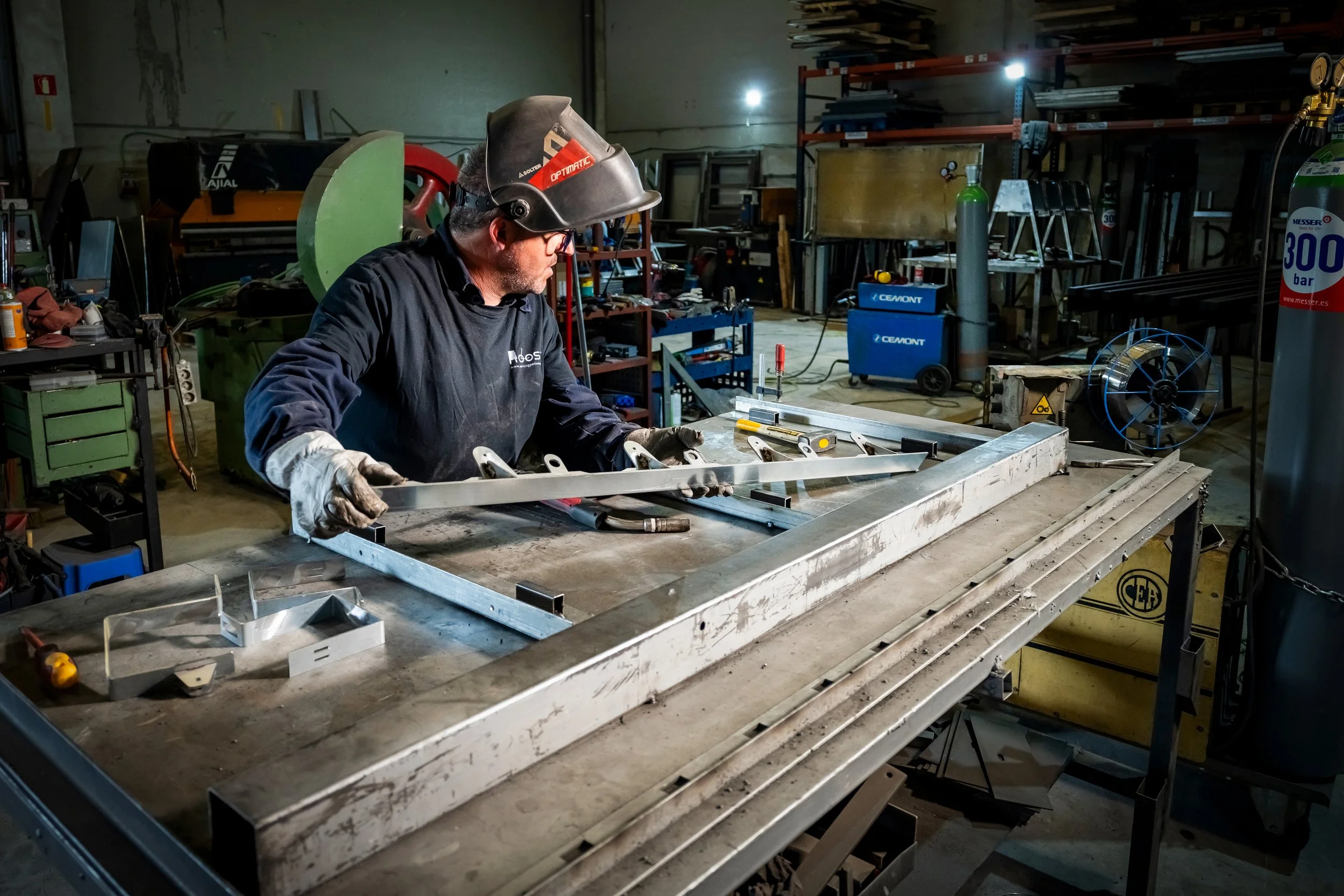 Un trabajador en un taller de metalurgia, usando equipo de protección, inspecciona una estructura metálica de Aparcapatinete en una mesa de trabajo rodeado de herramientas y maquinaria.