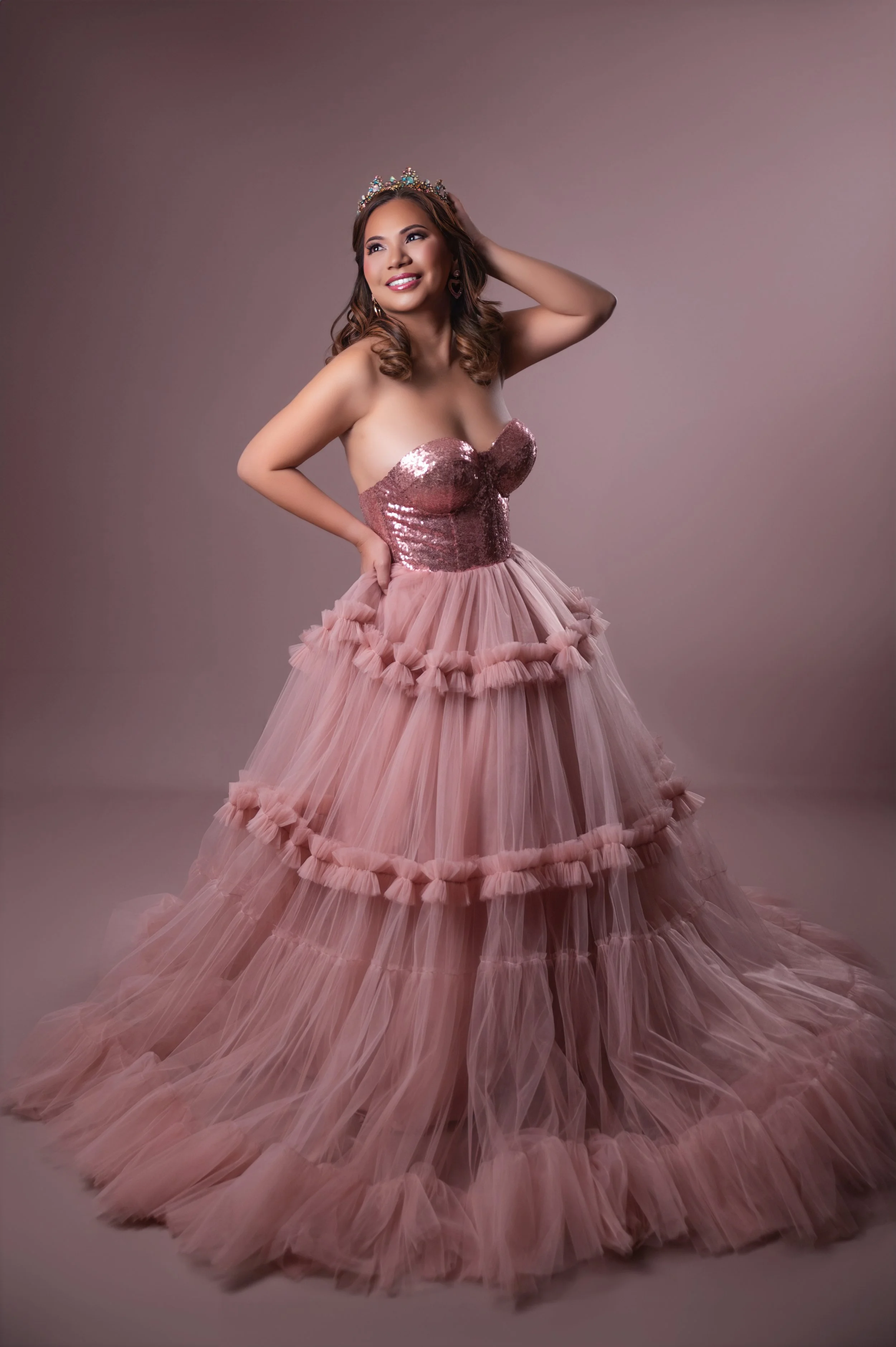 Woman in a sparkling pink gown and tiara poses joyfully against a soft pink background, exuding elegance and confidence.