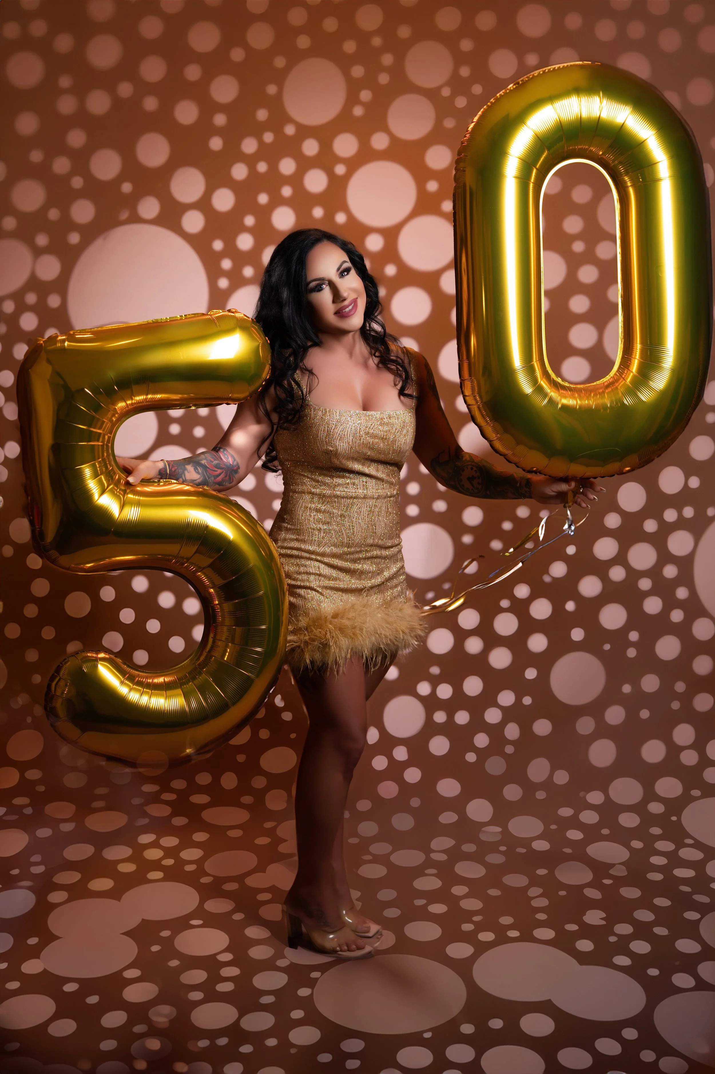 A woman in a glittery gold dress holds golden balloons shaped as the number 50. She's smiling against a polka dot background, conveying celebration.