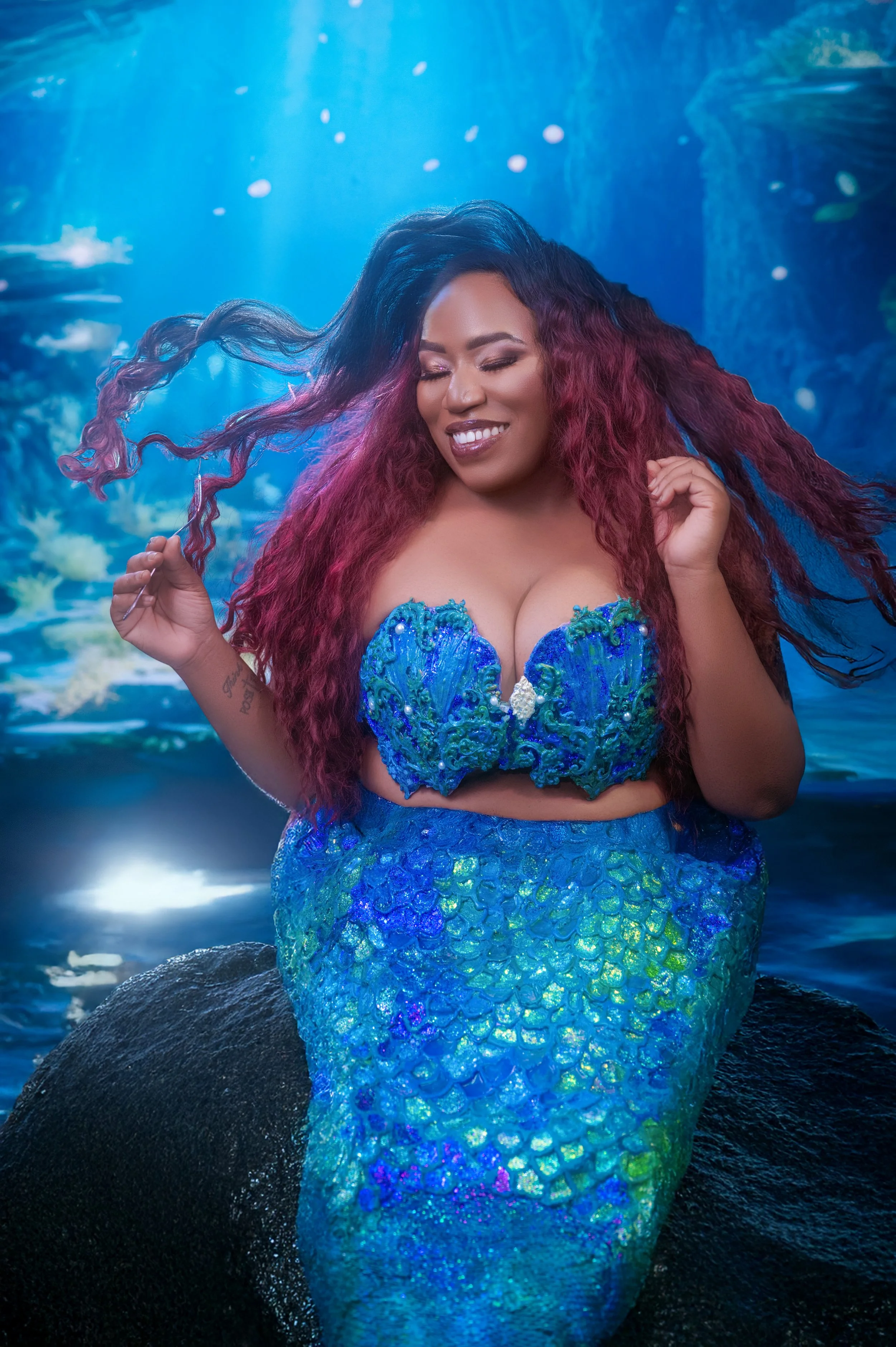A smiling woman with long, flowing red hair poses as a mermaid in a shimmering blue-green tail and top. The background is an enchanting underwater scene.