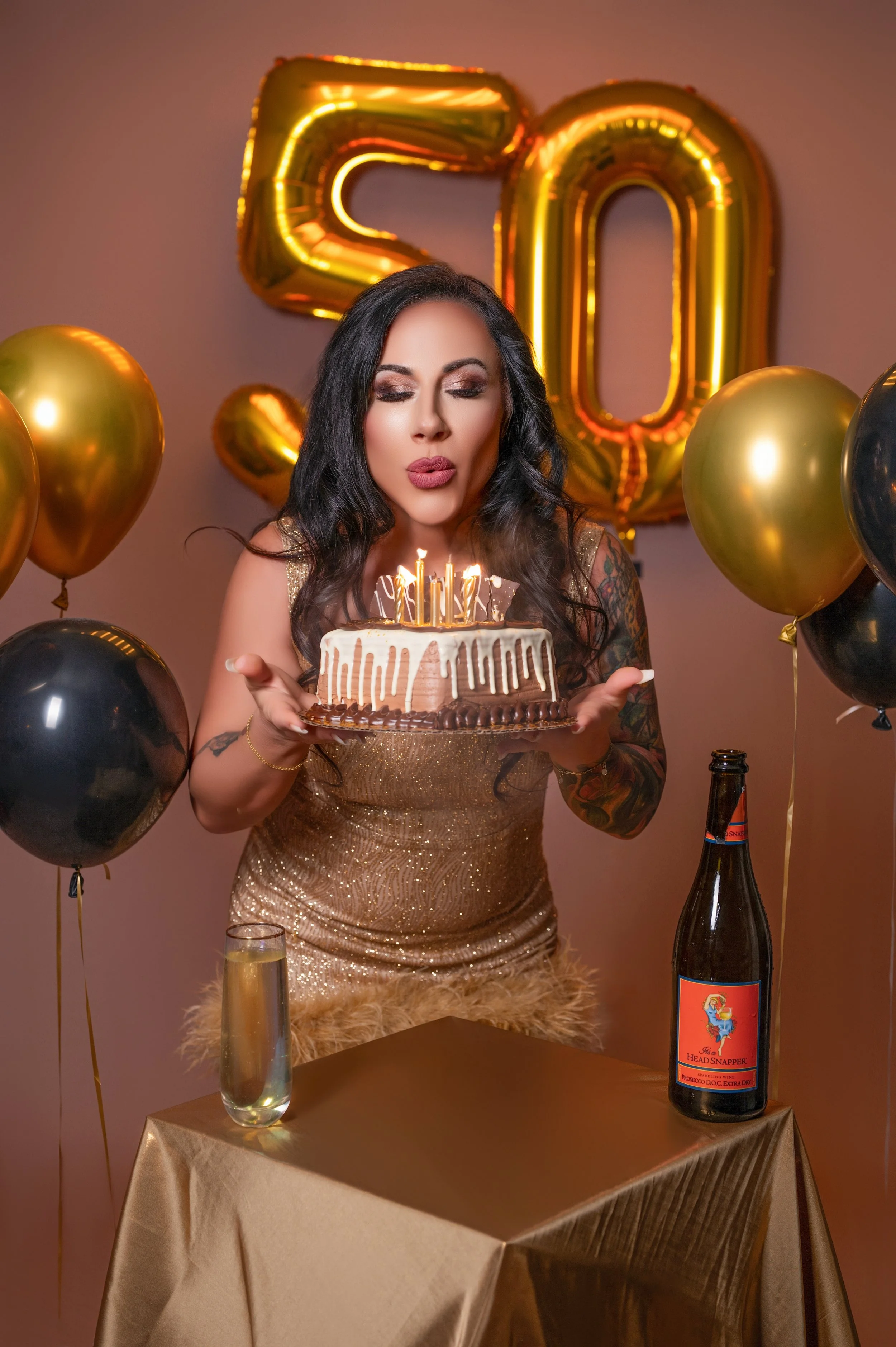 Woman in a sparkling gold dress blows out candles on a cake. Golden "50" balloons and black and gold balloons surround her, creating a festive atmosphere.