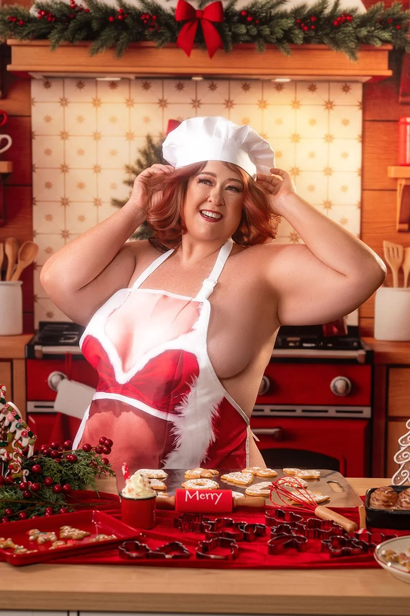 A laughing woman in a white chef's hat and red Santa apron stands at a kitchen counter covered in Christmas cookies and cookie cutters.
