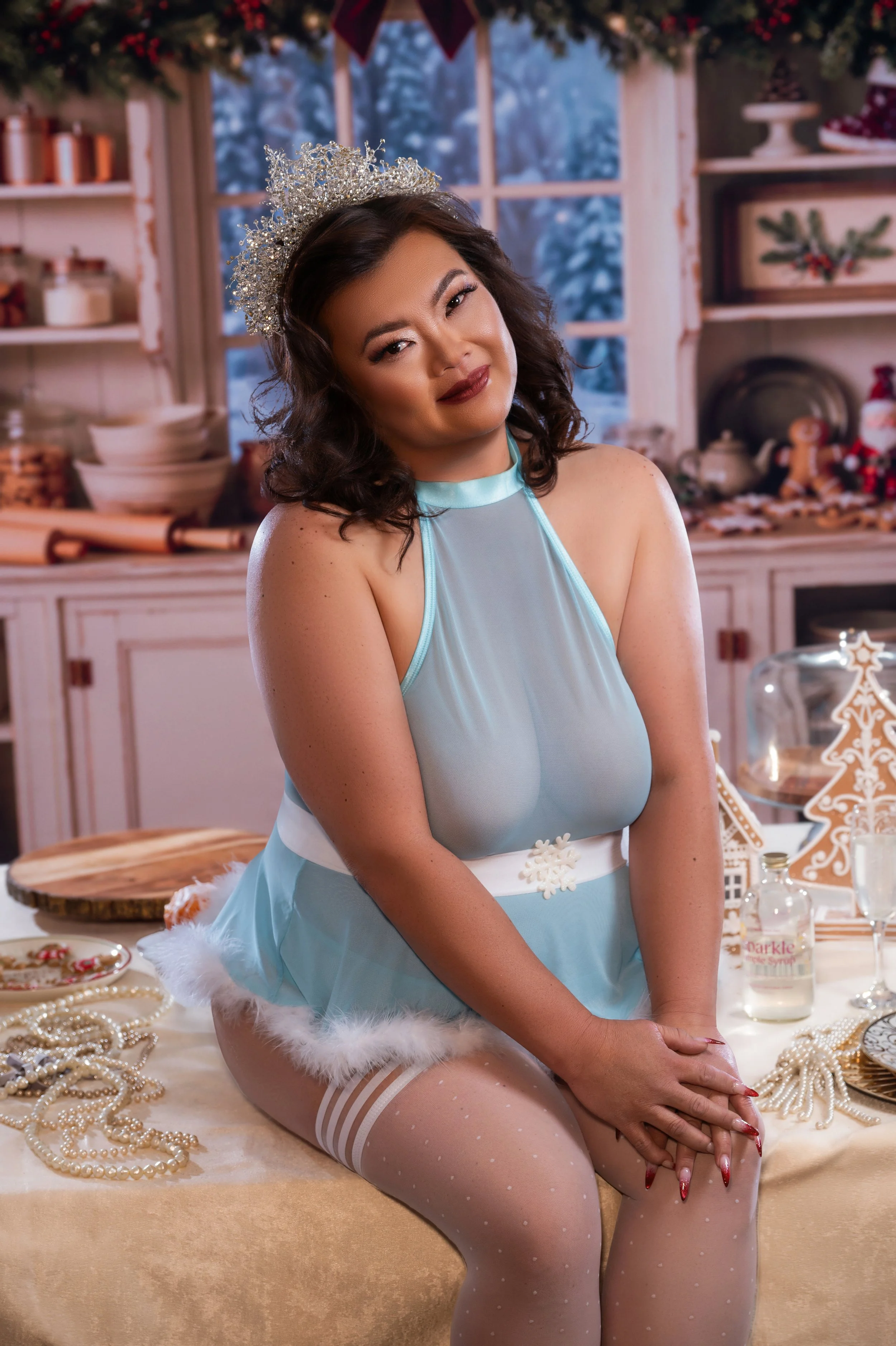 A smiling woman in a sparkly tiara and pale blue babydoll with white trim sits on a table in a cozy holiday kitchen set with snowy windows.