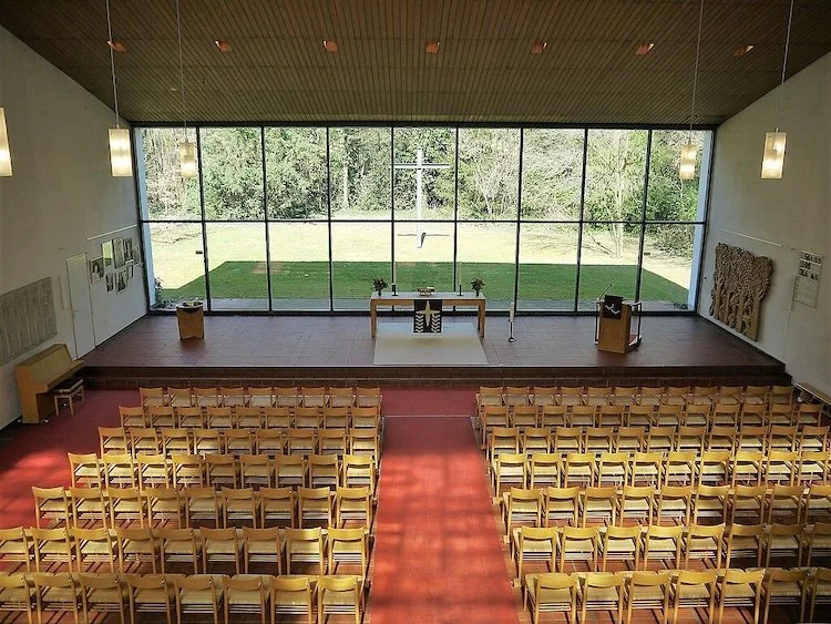 Church sanctuary with wooden chairs facing altar and a glass wall transparent to woodlands