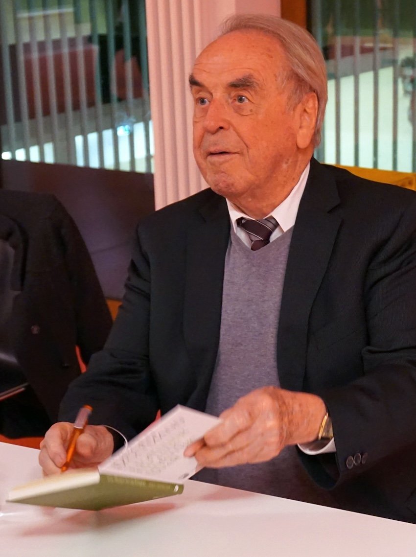 Seated man signing a book and looking straight ahead