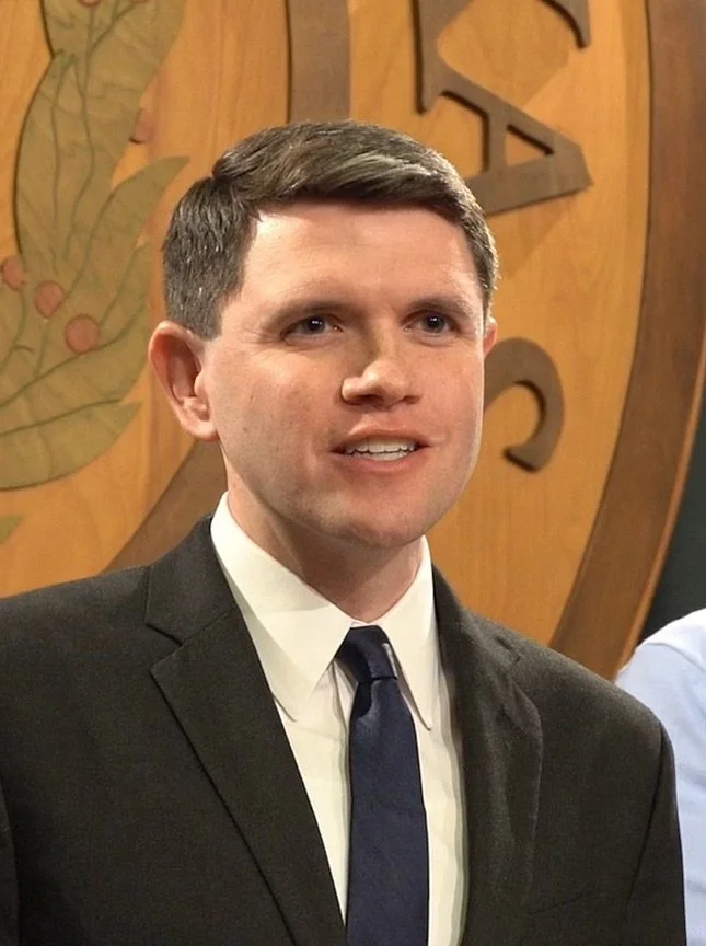 Man in black coat and tie speaking at news conference