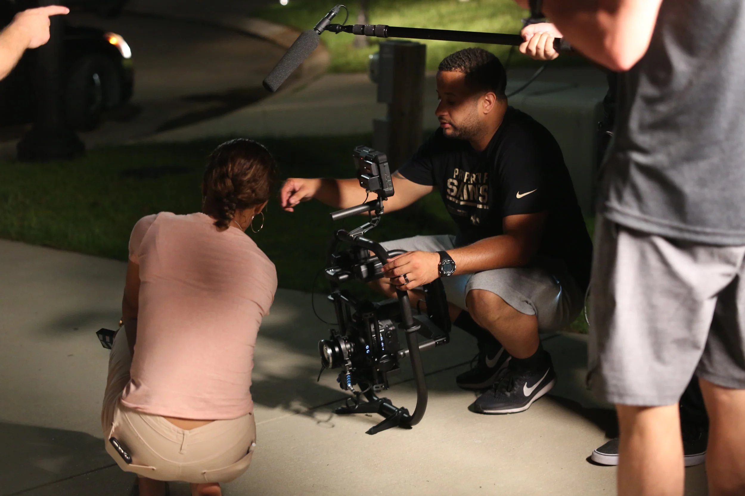 Filmmaking crew filming a scene outdoors at night, with the camera focused on a woman kneeling on the sidewalk.