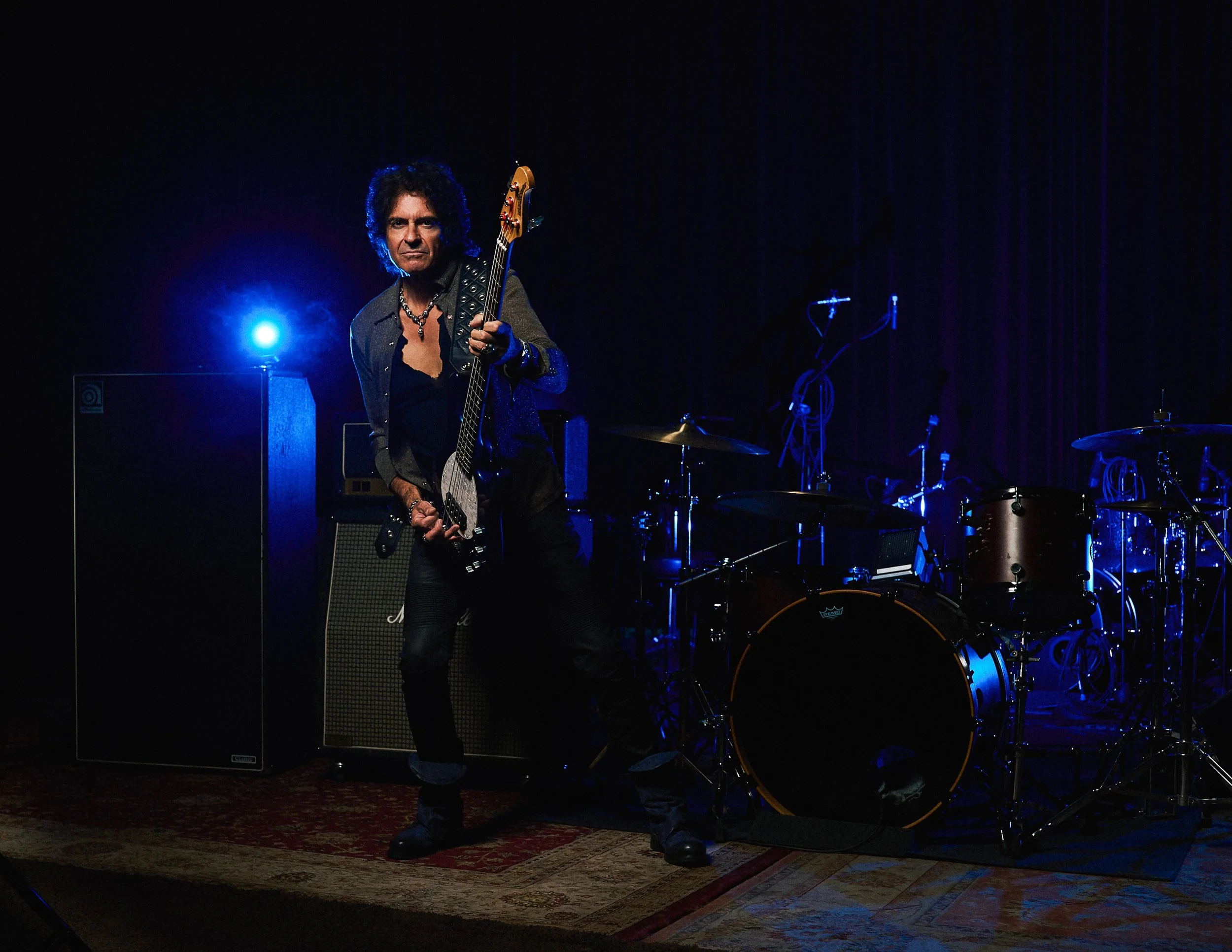 Professional full body portrait of legendary rock bassist Phil Soussan holding his bass guitar in a performance-style studio environment.