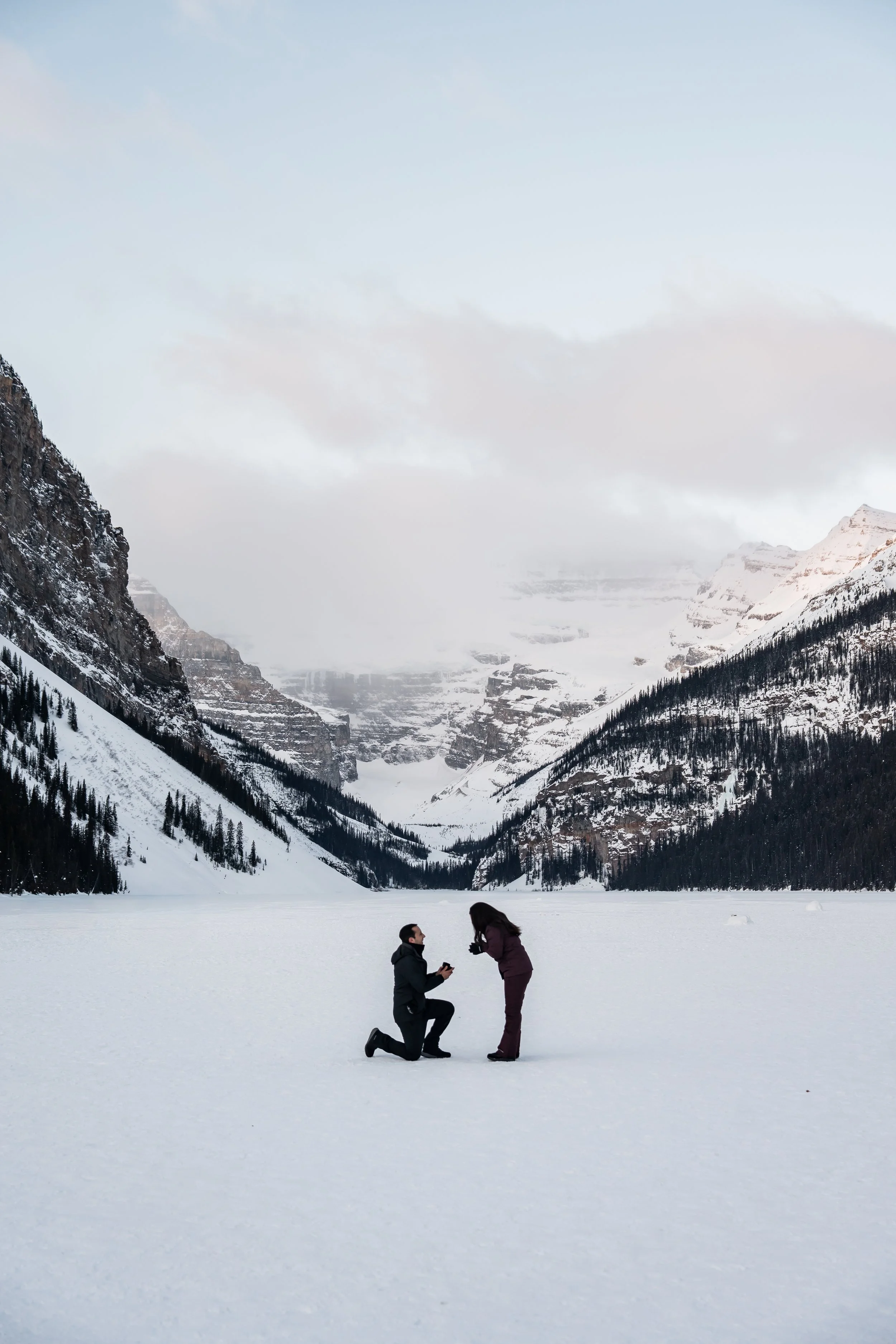 A snow-covered landscape with mountains in the background and two people in the foreground. One person is kneeling on one knee offering a ring, and the other is standing with hands over their face, possibly surprised or emotional.