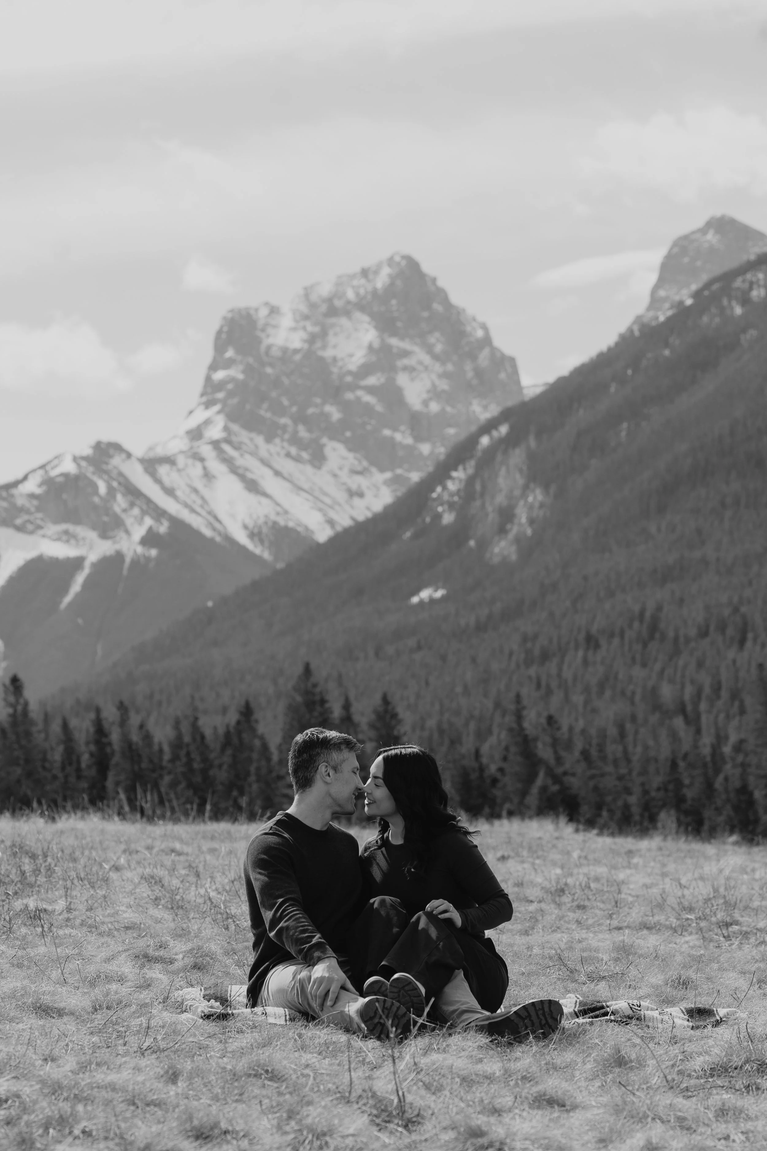 A black-and-white photo of a young couple sitting on the grass in a field, with mountains in the background and trees lining the horizon. The couple is facing each other closely.