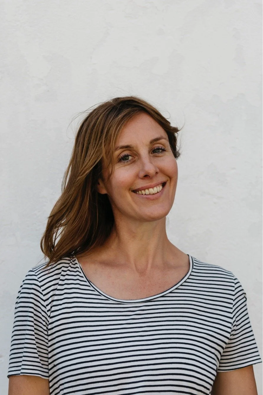 A woman with light brown hair in a striped shirt smiling against a white wall.