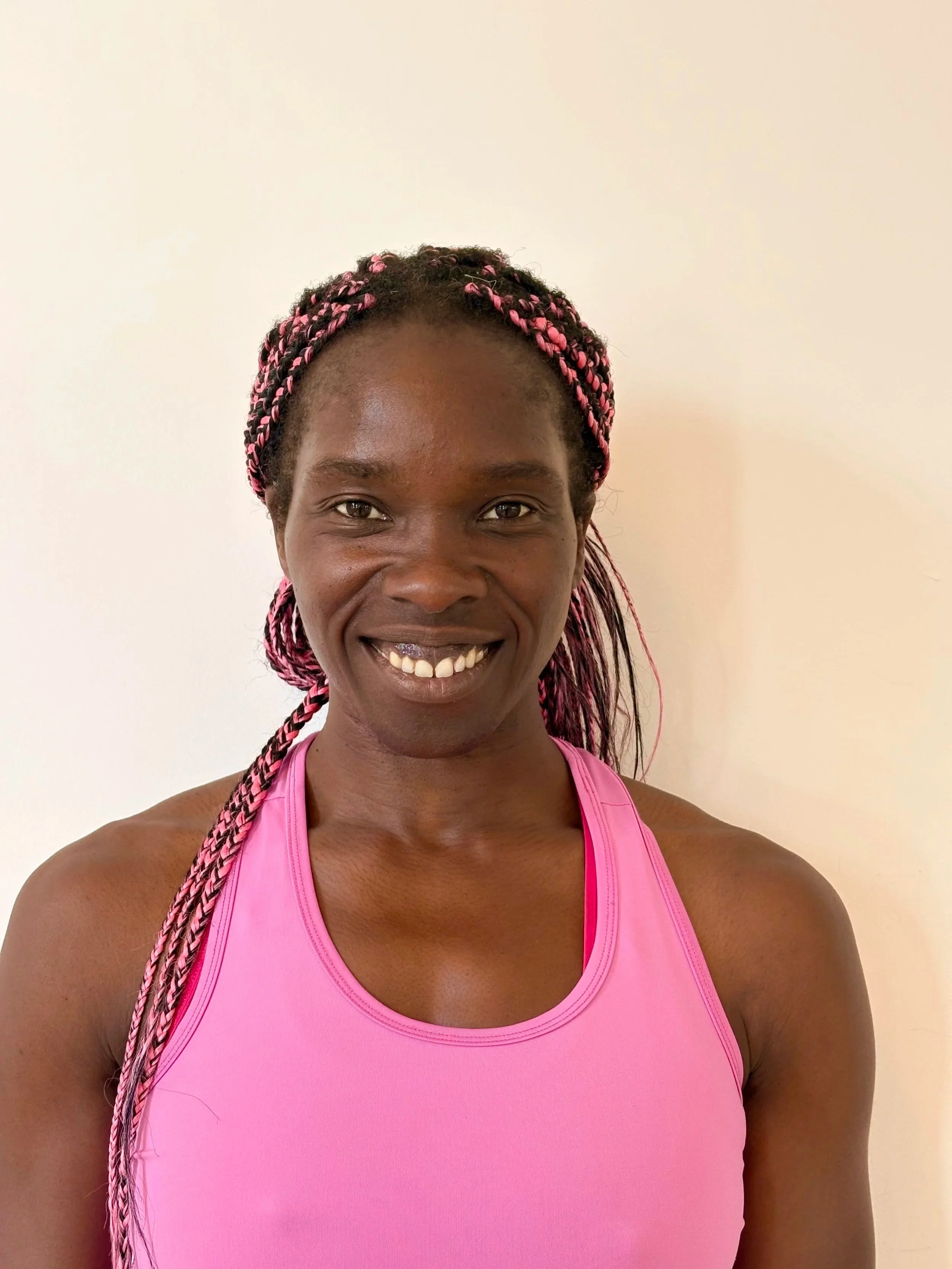 A smiling woman with braided hair wearing a pink workout top against a plain white wall.