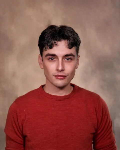 A young man with short dark hair wearing a red sweater against a neutral background.