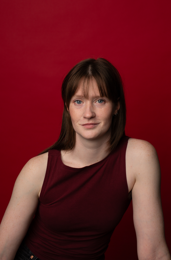 Portrait of a young woman with brown hair and blue eyes, wearing a sleeveless maroon top, against a red background.