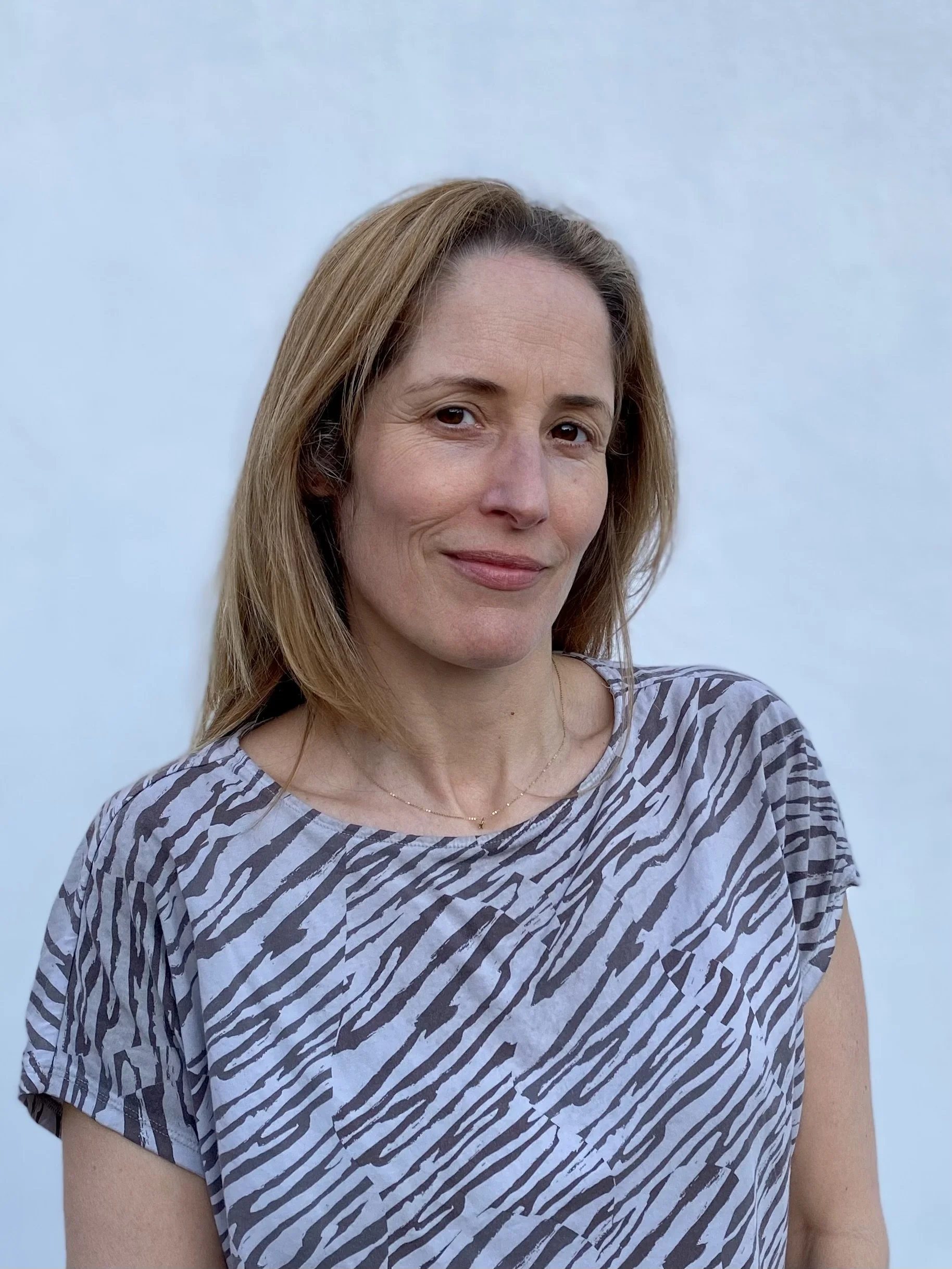 A woman with shoulder-length light brown hair smiling slightly, wearing a zebra-patterned shirt with a rounded neckline, standing against a plain light blue background.