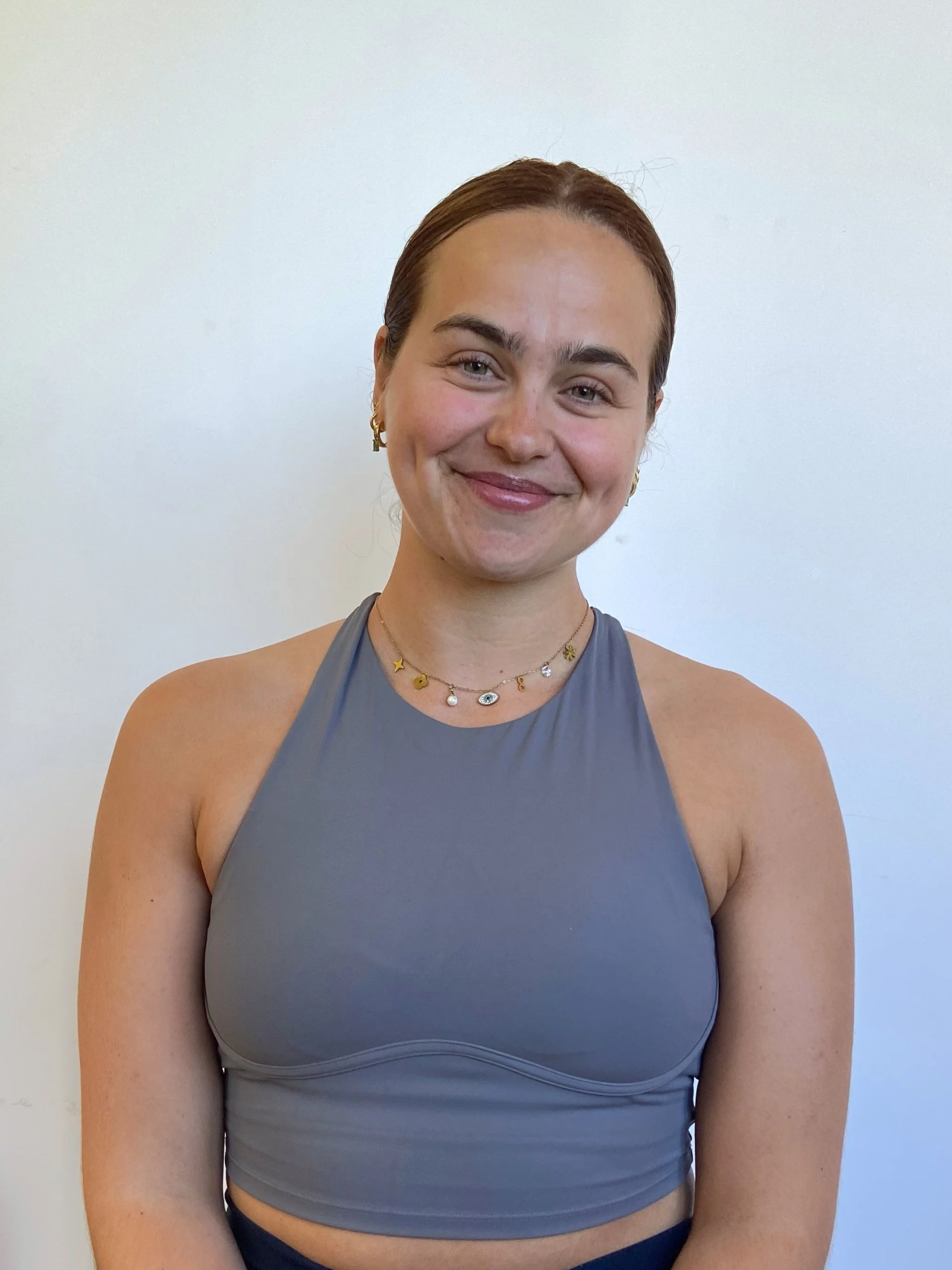 A young woman with short brown hair, wearing a sleeveless gray top and jewelry, smiling against a plain white background.