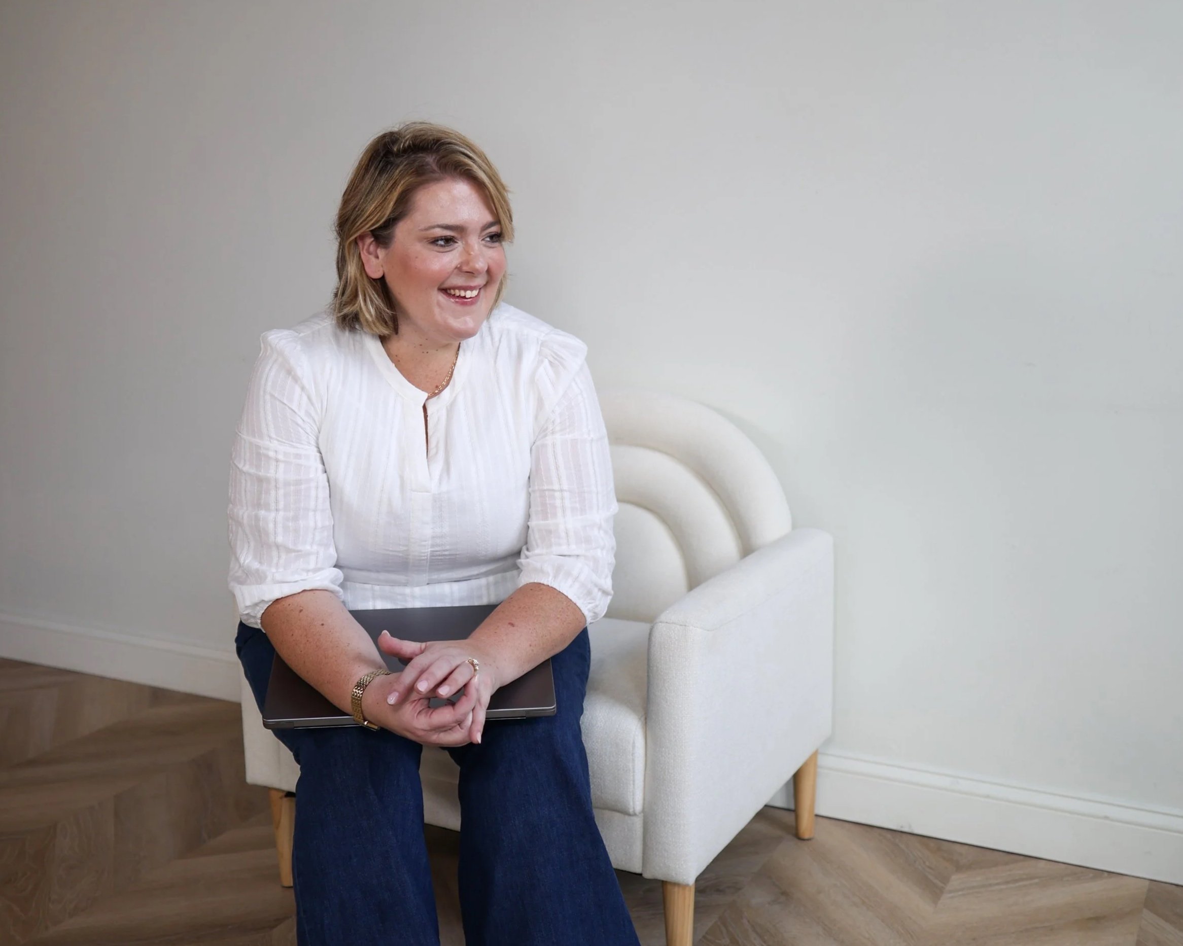 A woman with blonde hair, wearing a white blouse and blue jeans, sitting on a cream-colored armchair, holding a closed laptop, smiling, in a room with wooden flooring and white walls.