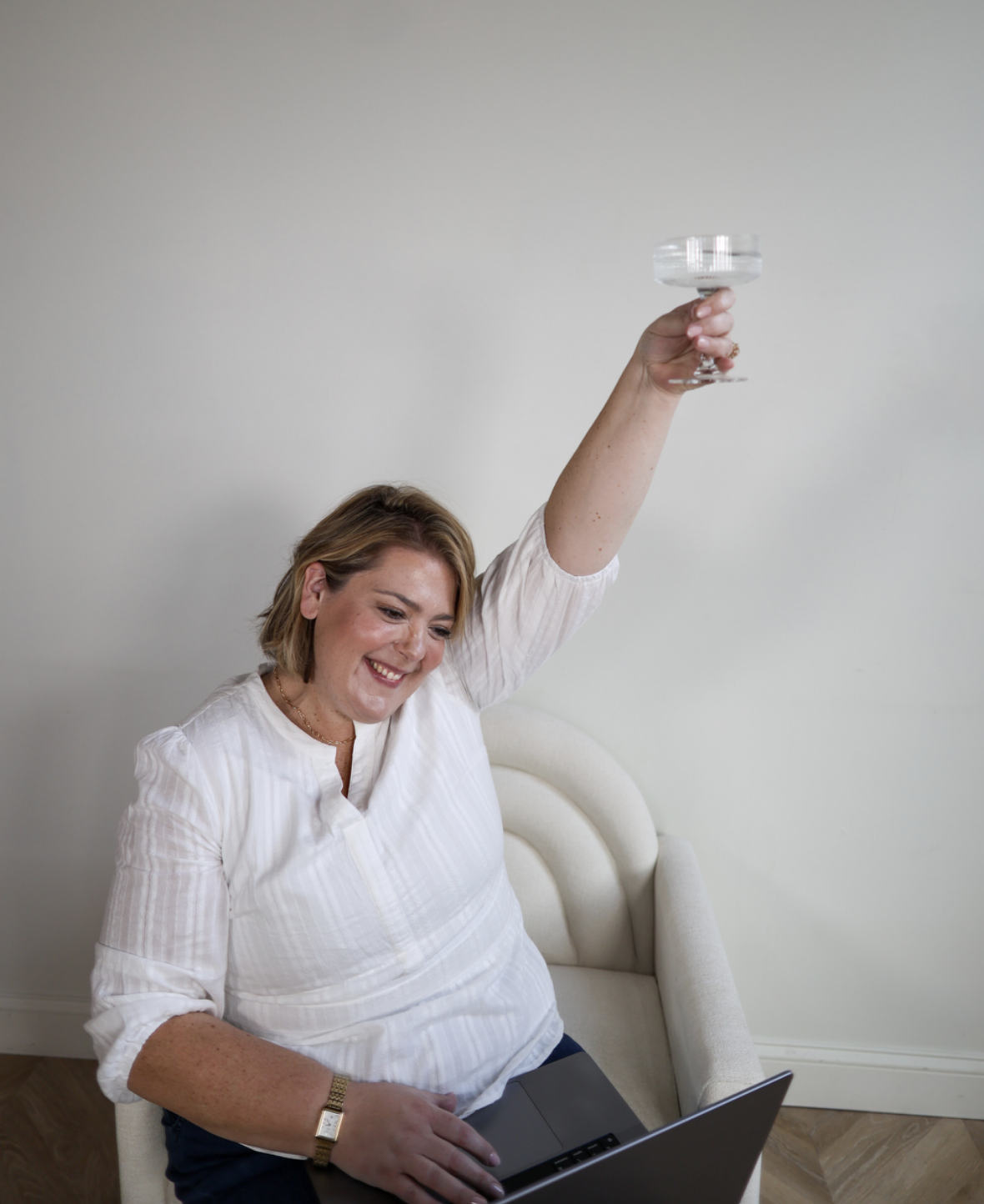 A woman smiling and raising a glass in a toast while sitting on a chair with a laptop on her lap.