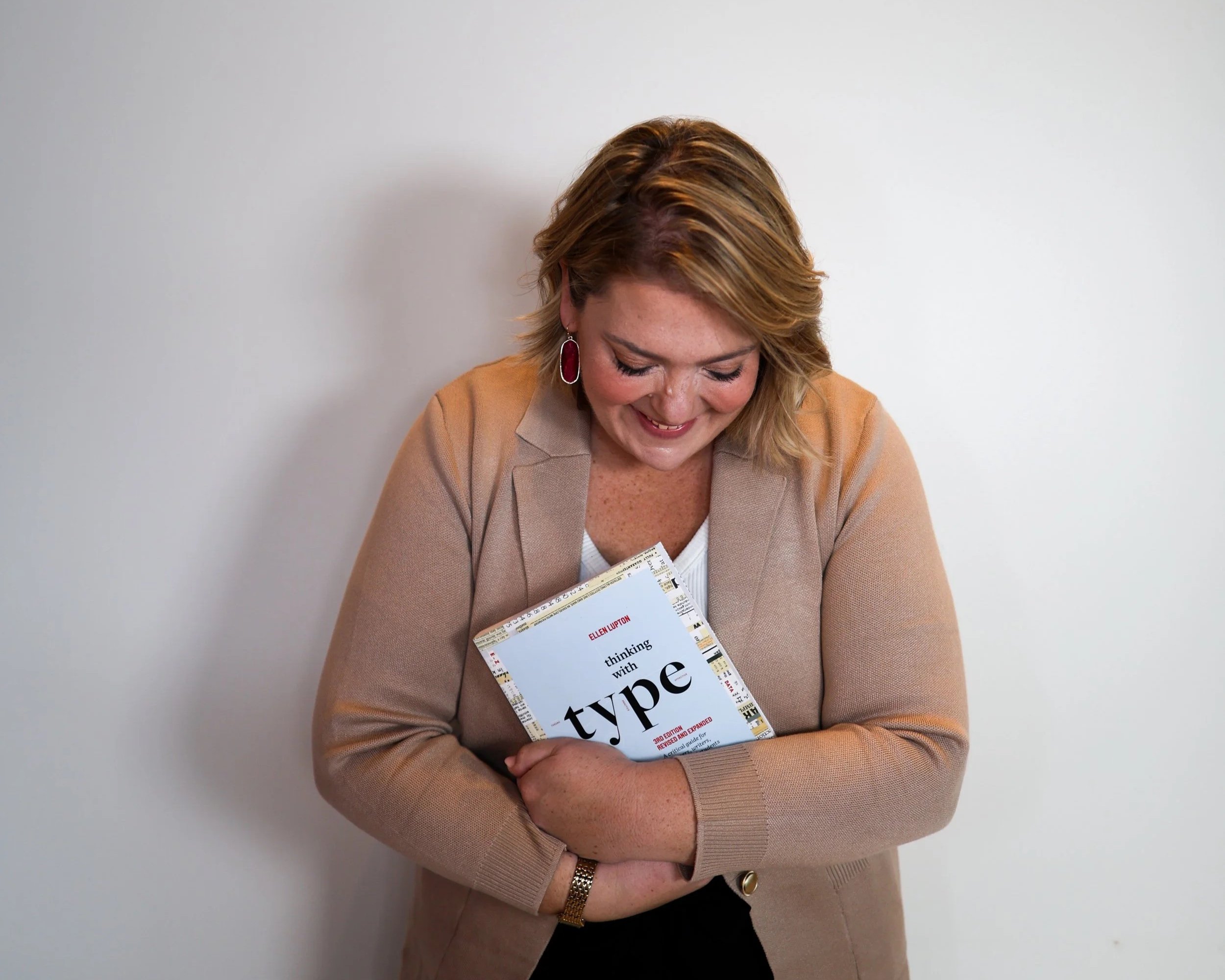 A woman with shoulder-length blonde hair, wearing a beige blazer and a white top, is smiling while holding a book titled 'Thinking with Type' by Ellen Lupton, against a plain white wall.