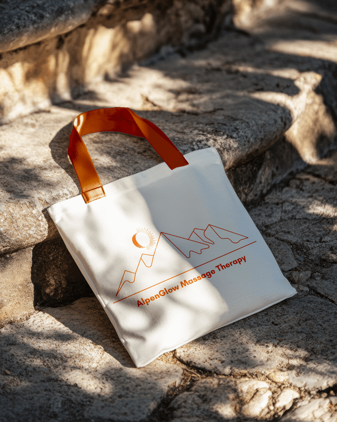 Branded tote bag for AlpenGlow Massage Therapy placed on outdoor stone steps, showing brand applied to physical product