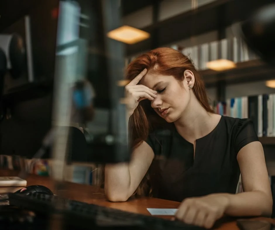 Woman sitting at a desk in her office with her hand on her forehead has the appearance of being frustrated and overwhelmed  with workload