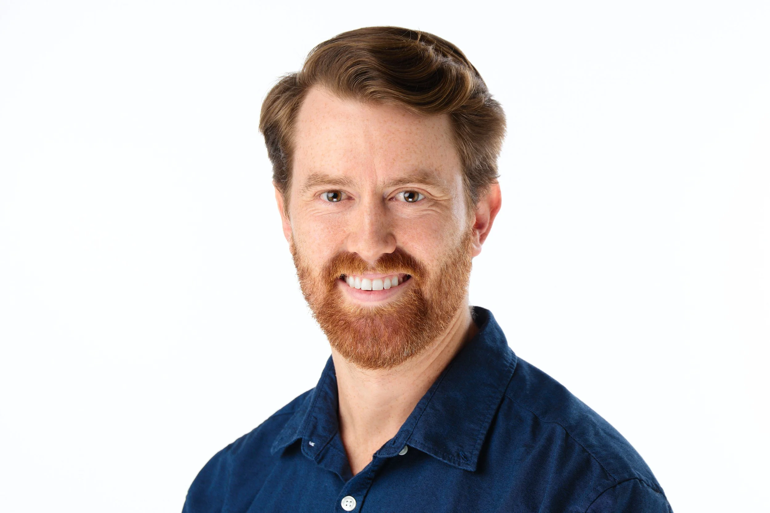 A man with red hair, a beard, and mustache, wearing a blue shirt, smiling against a plain white background.