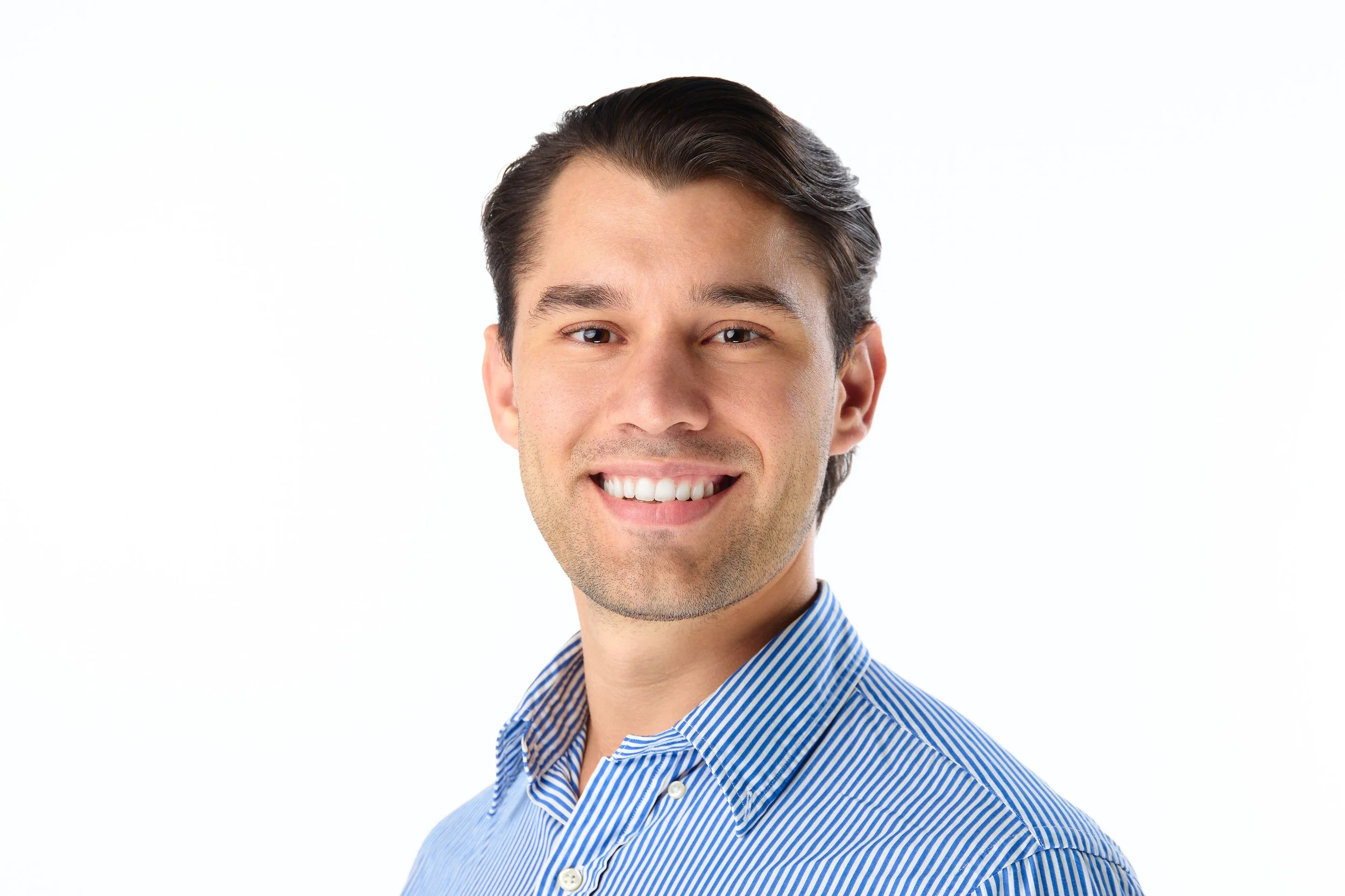 A young man with dark hair, wearing a blue and white striped shirt, smiling against a plain white background.
