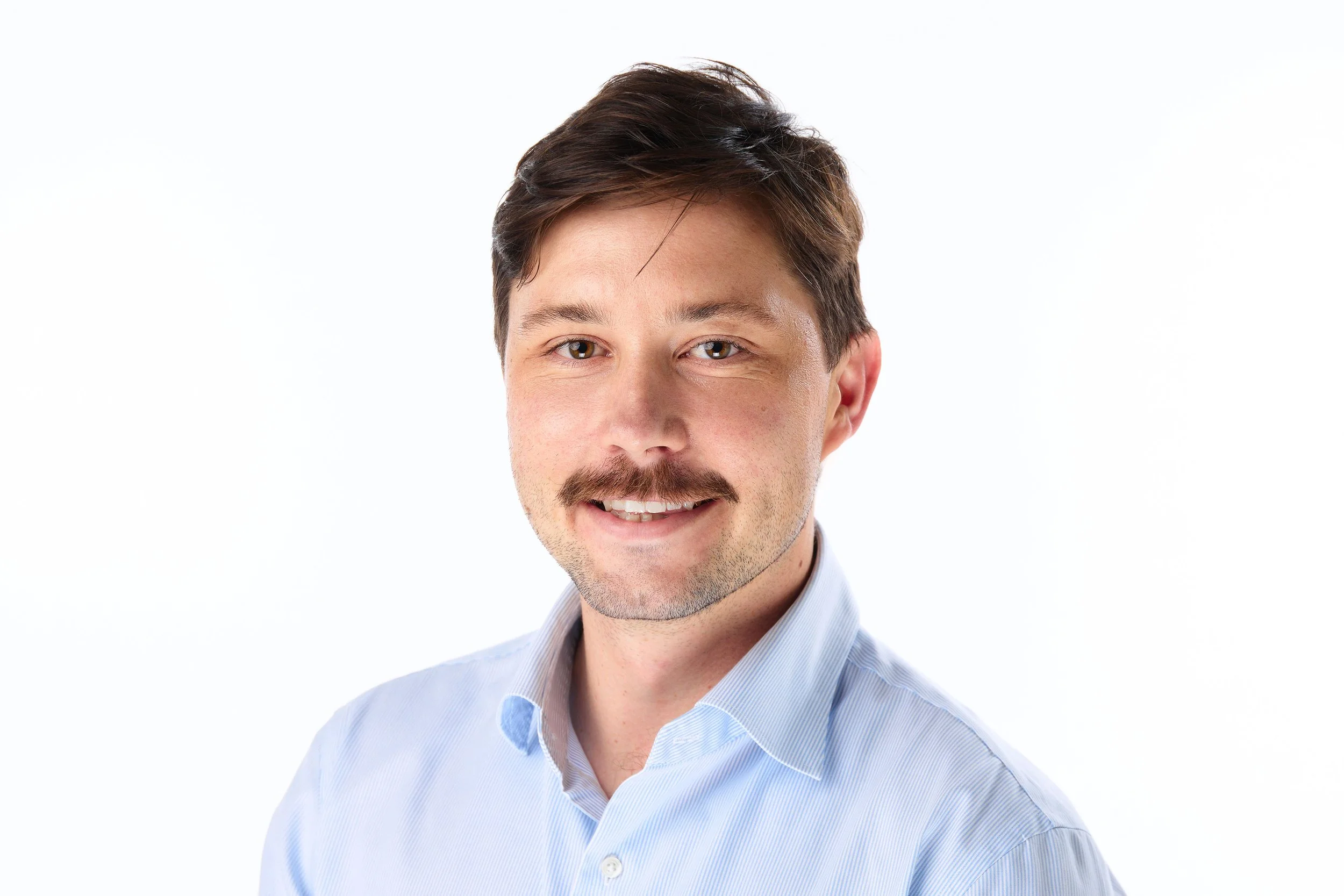 Head and shoulders of a smiling man with brown hair, wearing a light blue dress shirt, against a white background.