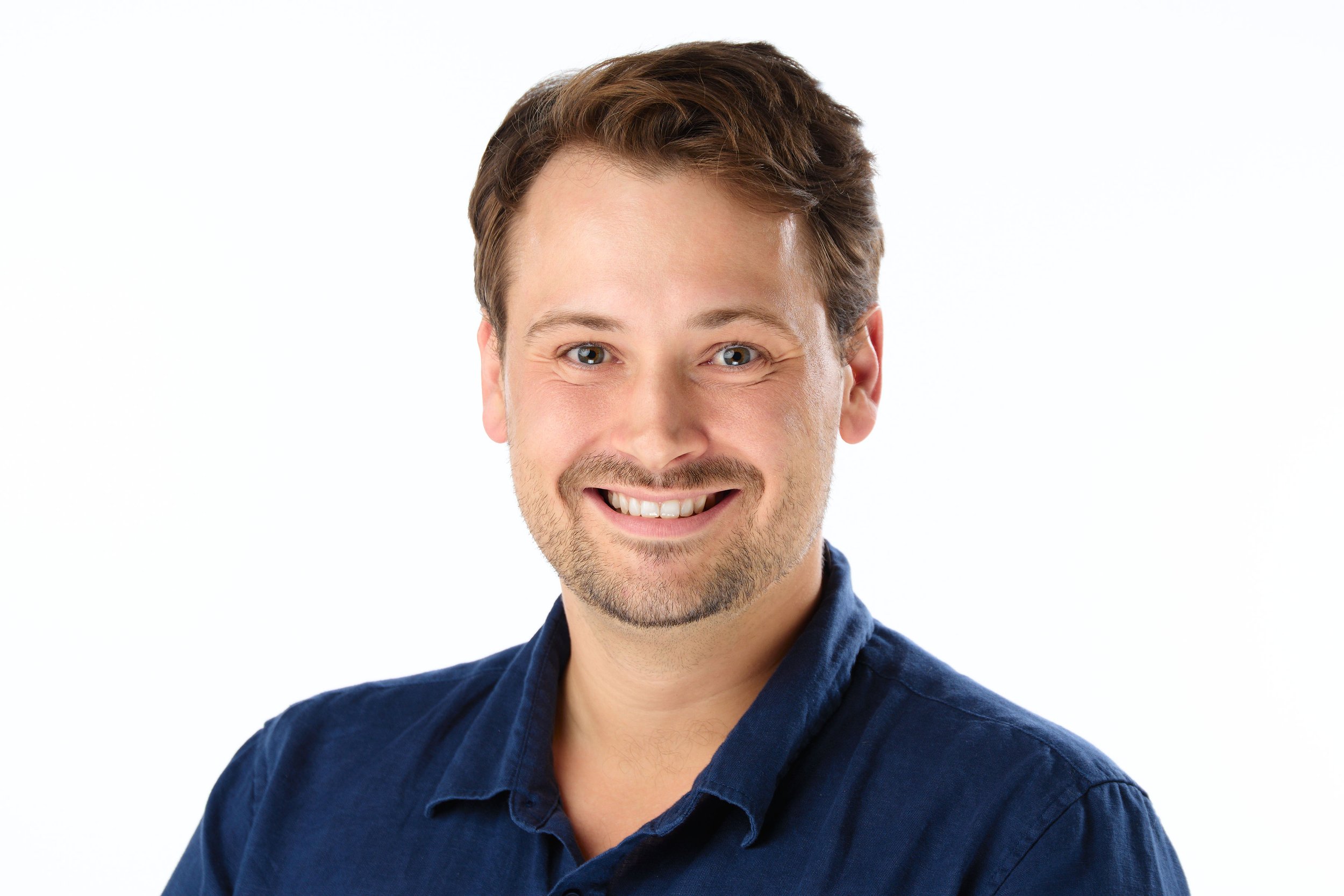 A smiling young man with brown hair, light facial hair, wearing a dark blue collared shirt, against a plain white background.