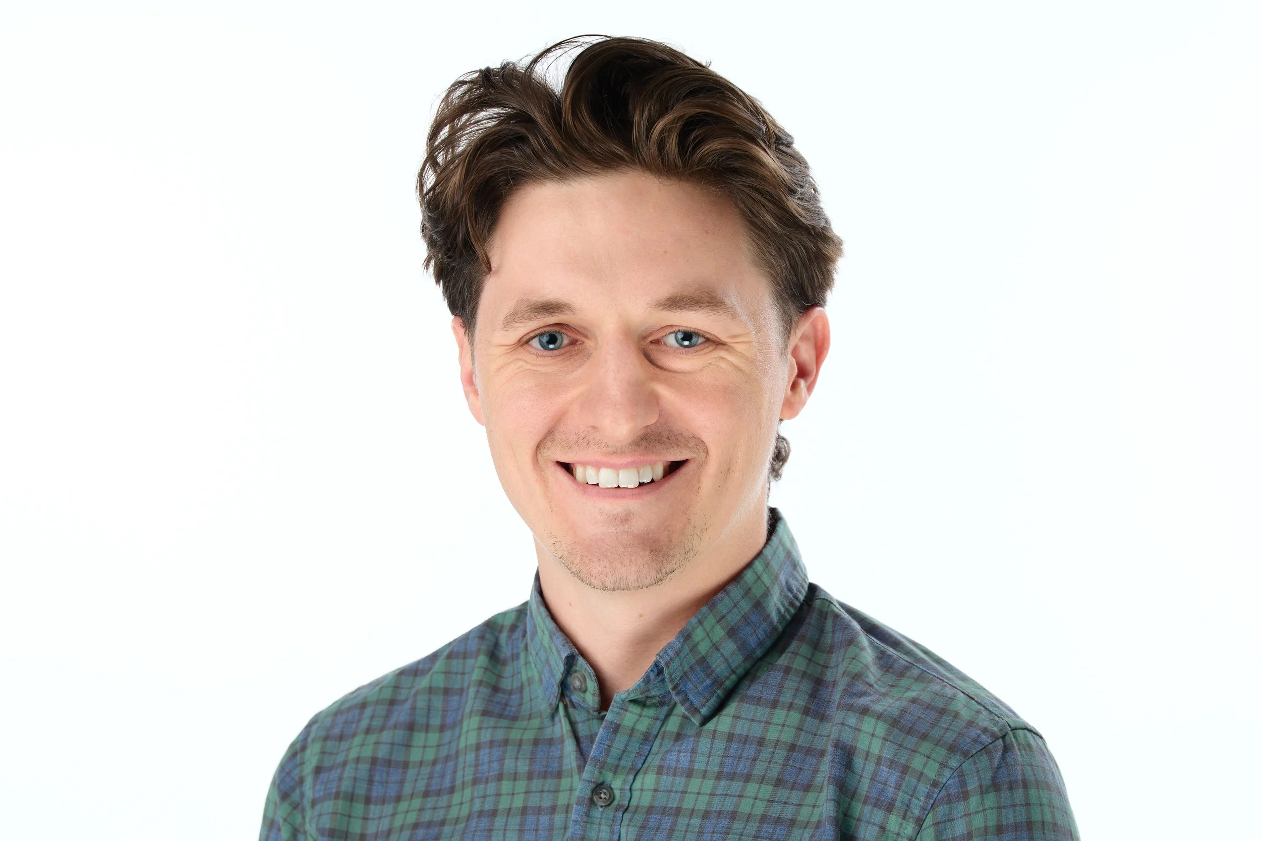 Young man with brown hair, blue eyes, wearing a green and blue plaid shirt, smiling, against a white background.