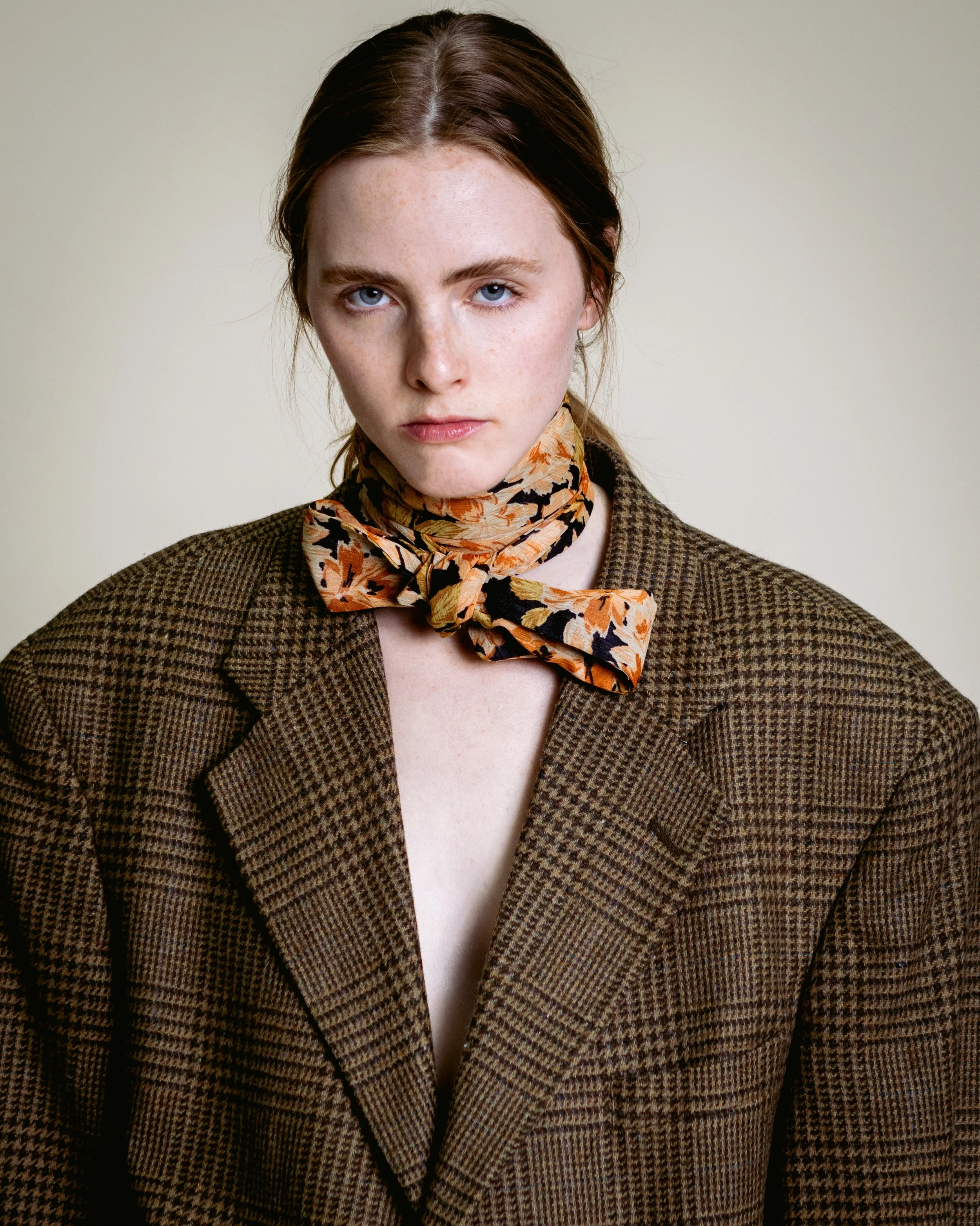A young woman with red hair, blue eyes, and freckles, wearing a brown checkered blazer and an orange patterned scarf tied around her neck, looking directly at the camera with a serious expression.