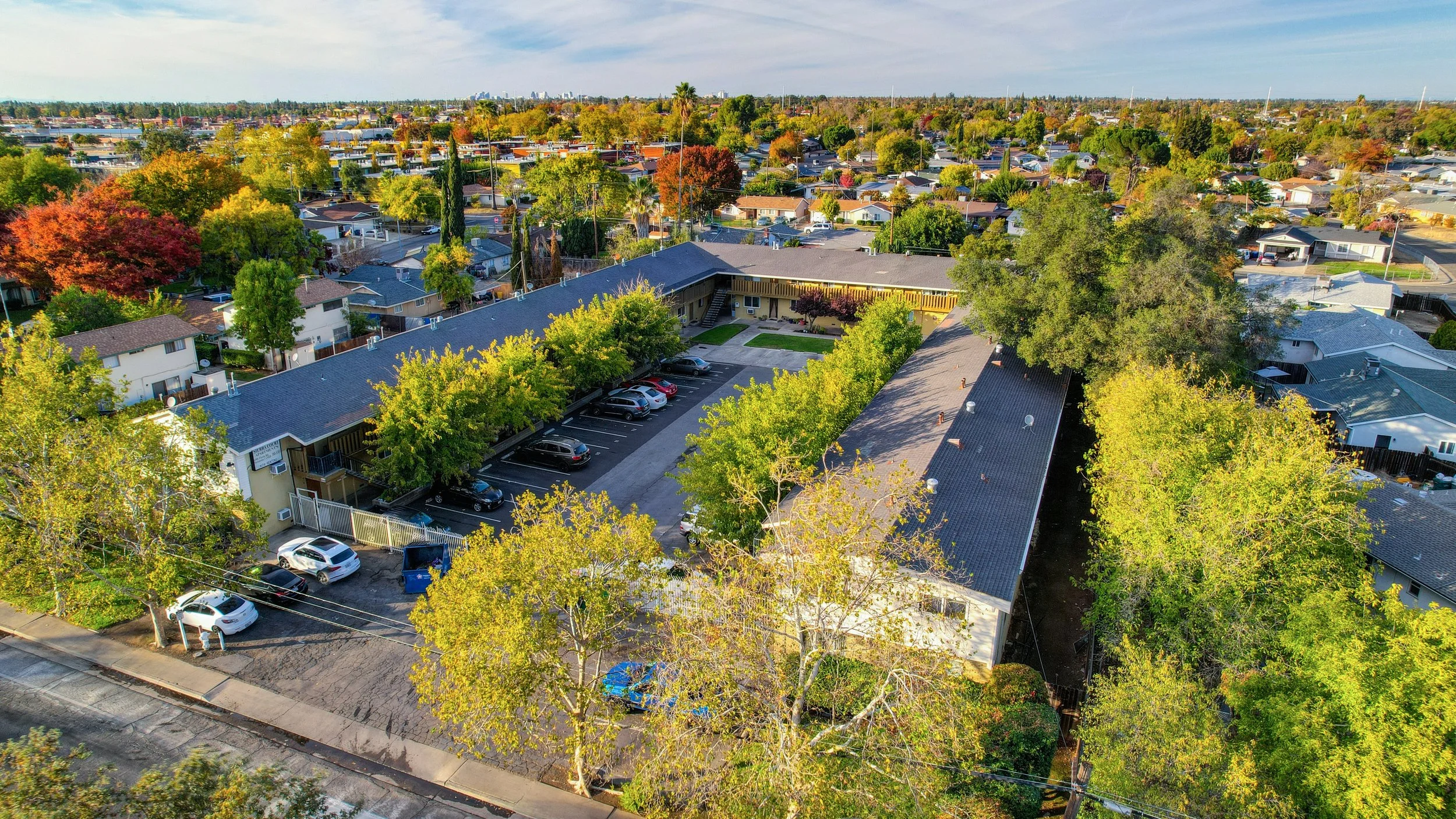 Aerial view of a residential neighborhood with single-family homes, trees with autumn foliage, a parking lot with cars, and a large building with a dark roof.