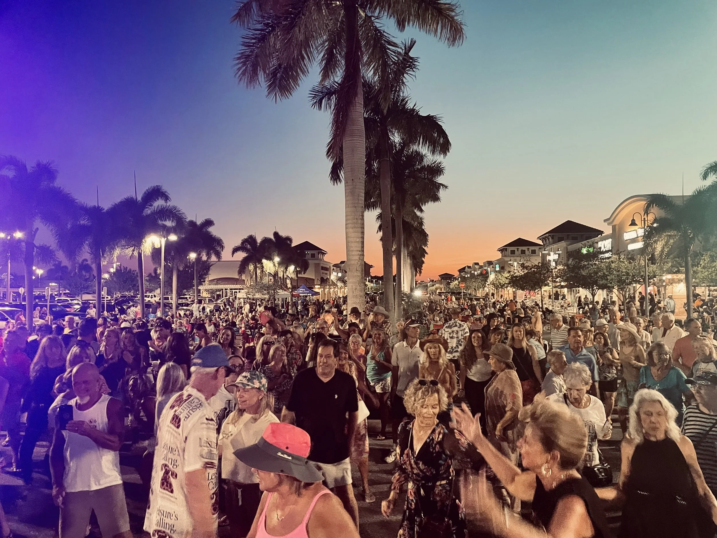 Large outdoor crowd at sunset with palm trees and shops in the background.
