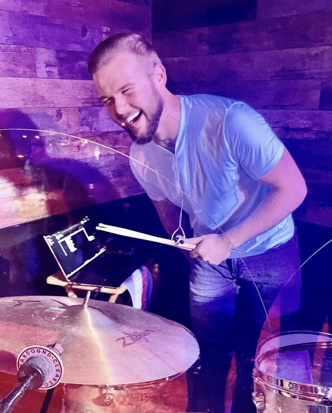 Man with headphones and drumsticks smiling behind a drum set with Zildjian cymbals and wood panel background.