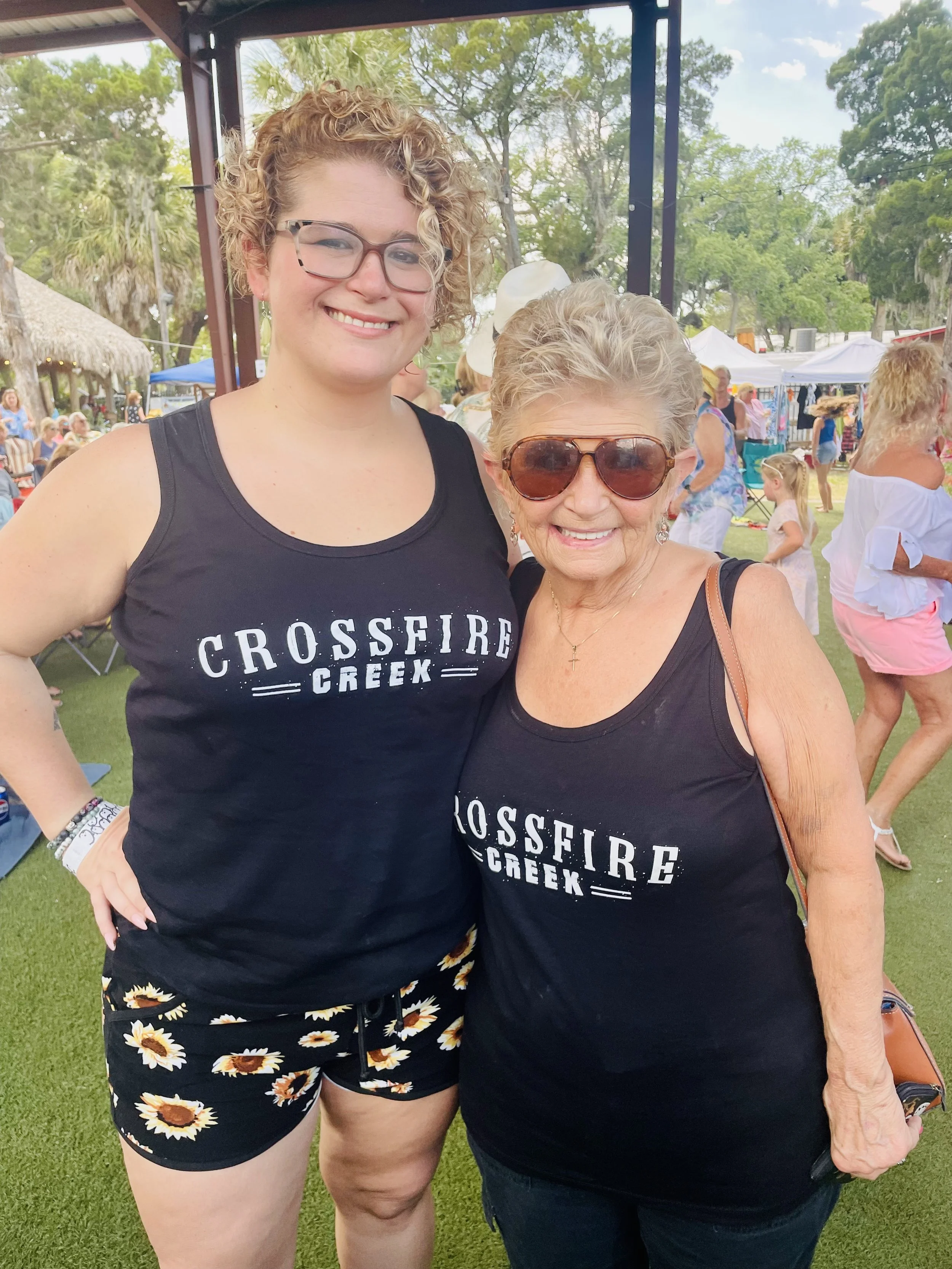 Two women smiling outdoors at an event, both wearing black tank tops with "Crossfire Creek" printed on them. The younger woman is wearing glasses and floral shorts, while the older woman is wearing sunglasses. People and tents are visible in the back