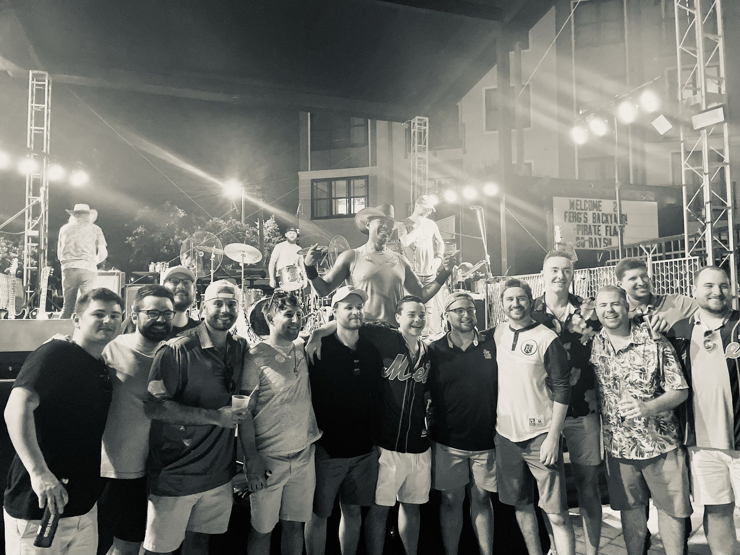 Group of men posing in front of a stage at a concert event, with musicians and instruments in the background under stage lights, in black and white.