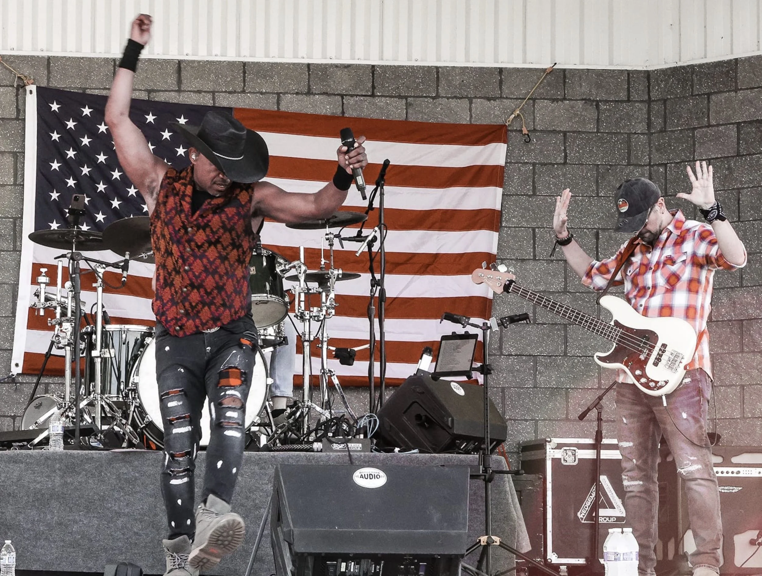 Country music band performing on stage in front of an American flag. The lead singer wears a cowboy hat and vest, while the bassist in a plaid shirt holds a guitar. Drums and other musical equipment are visible.