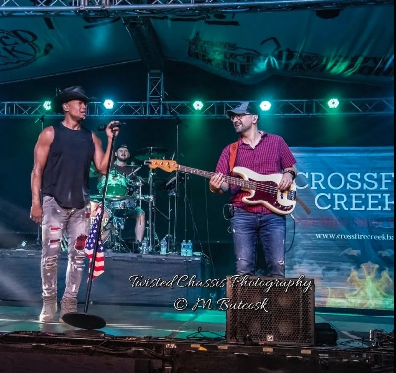 Musician performing on stage with a bassist and drummer, American flag attached to the microphone stand. Banner in the background reads 'Crossfire Creek.'